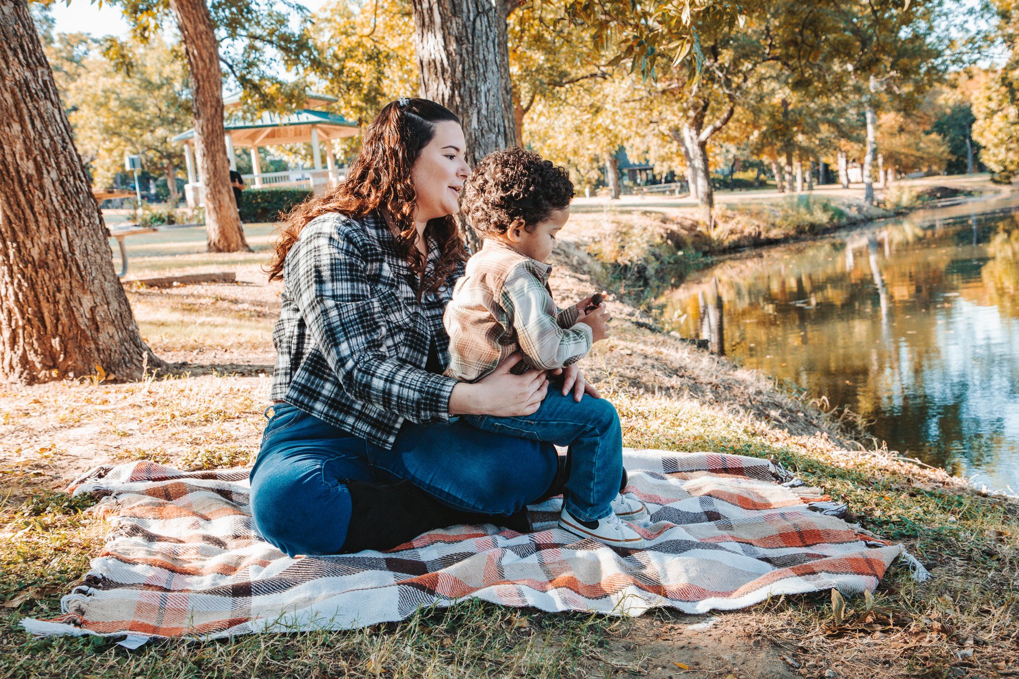 A woman and young boy sitting on a blanket by a river in a park during fall, with trees displaying autumn colors in the background.