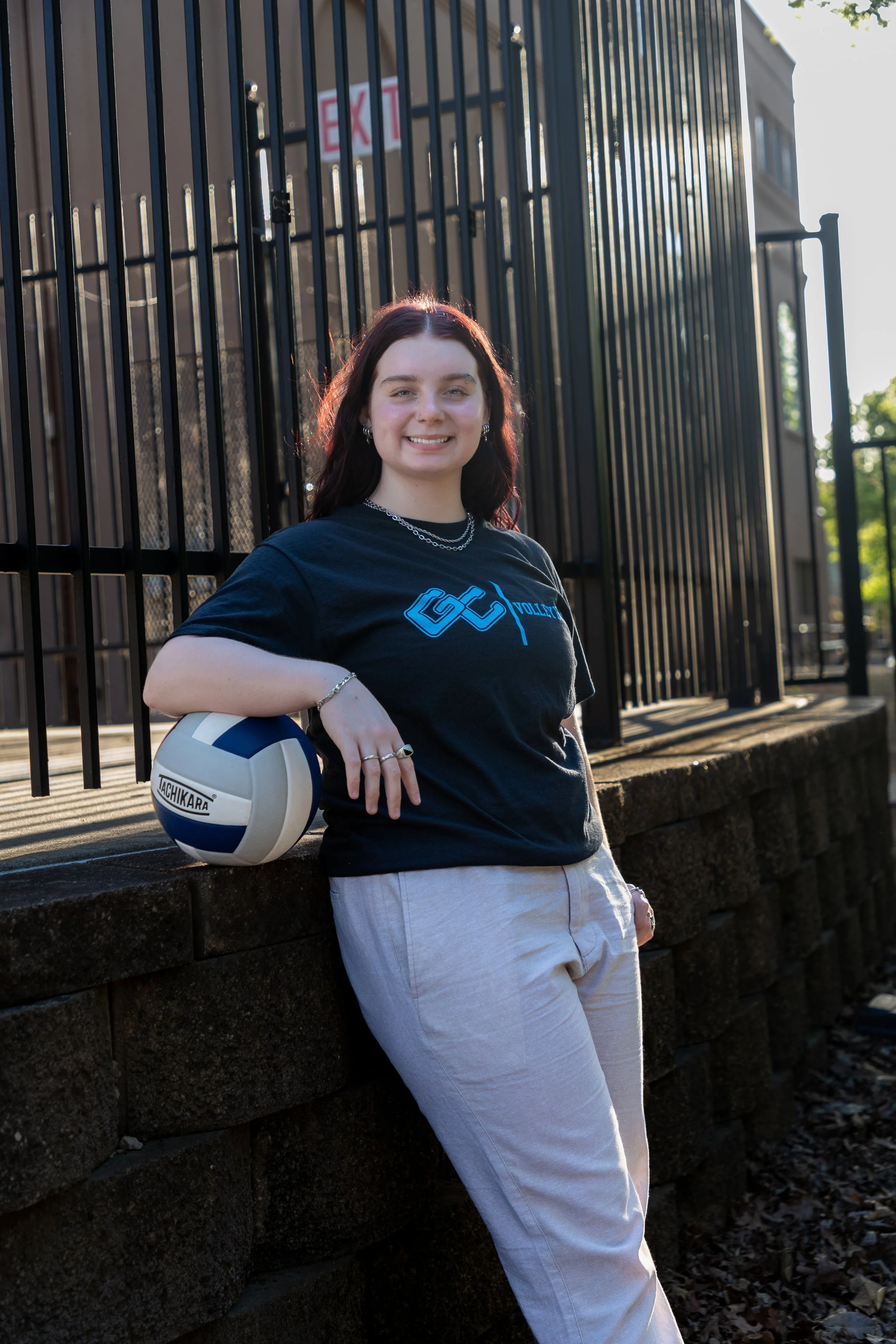 A young woman with dark red hair smiling and leaning against a stone wall outdoors, holding a volleyball in her right hand, wearing a black t-shirt and light-colored pants.