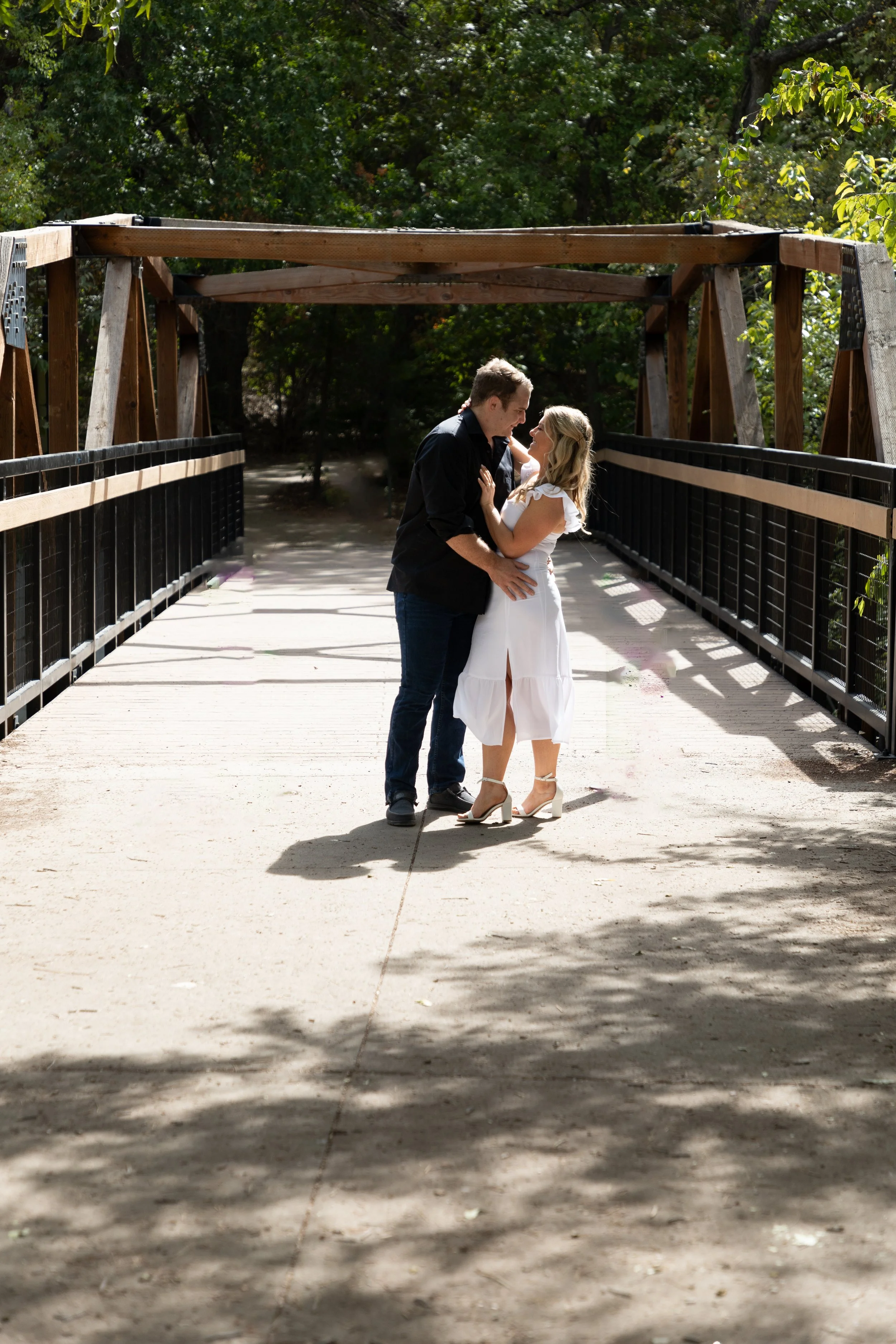 A couple dancing and embracing on a wooden bridge with trees and sunlight in the background.