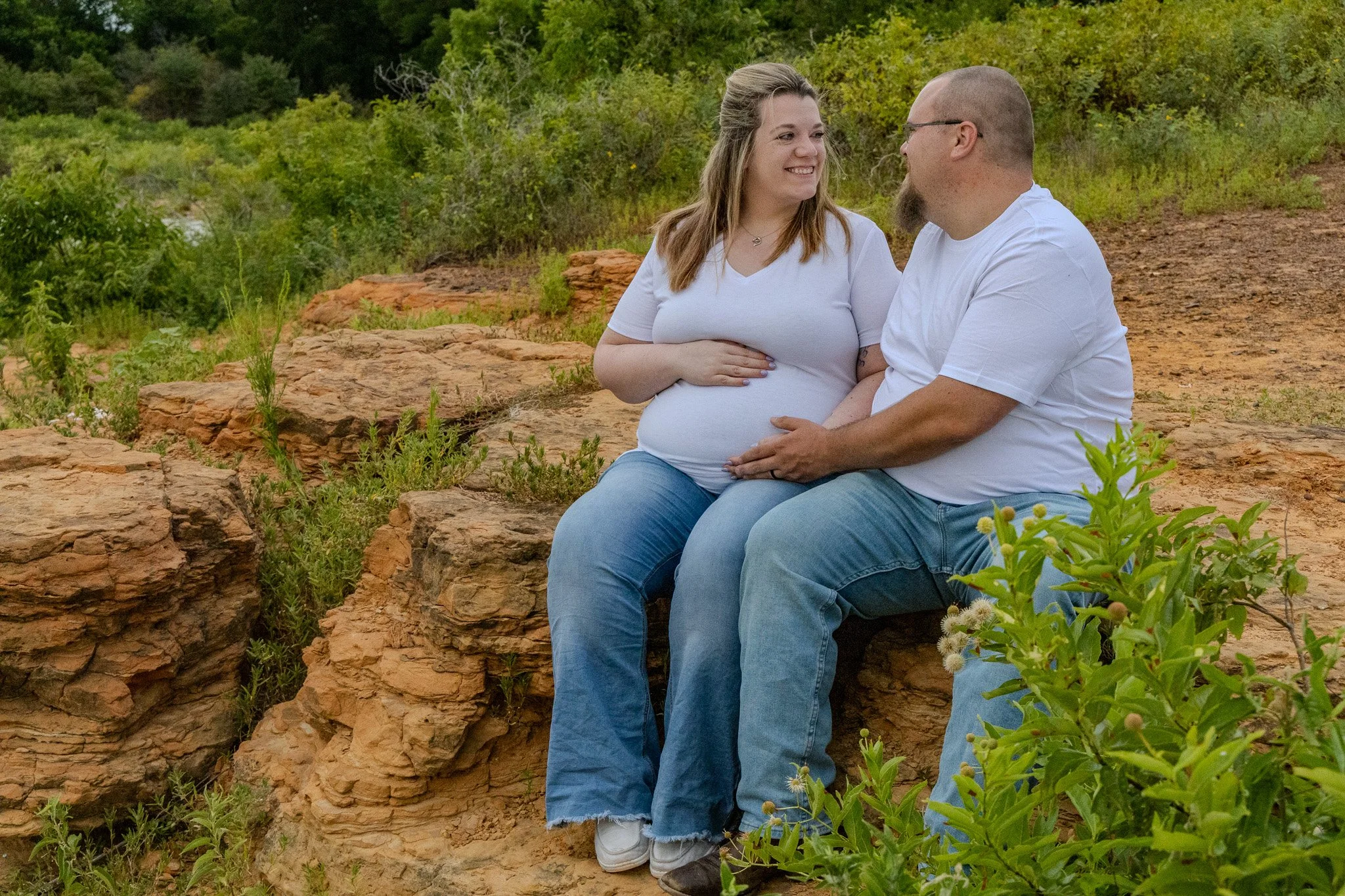 A smiling woman and man sitting on a rock in a natural setting, facing each other, with the woman touching her pregnant belly and a forested background.