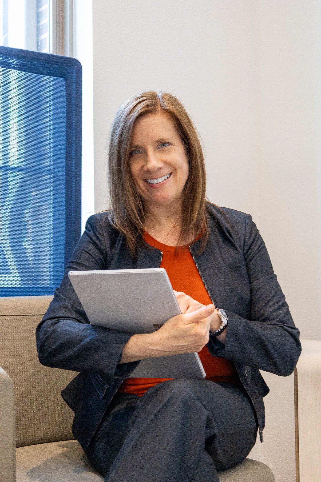 A smiling woman with shoulder-length brown hair, wearing a black jacket over an orange top, sitting on a beige chair holding a tablet, in an office environment.
