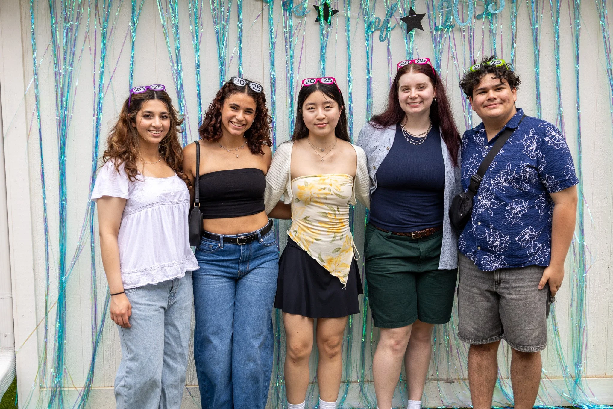 Group of five young adults celebrating graduation, wearing headbands indicating the year 2023, standing in front of colorful streamers and black stars backdrop.