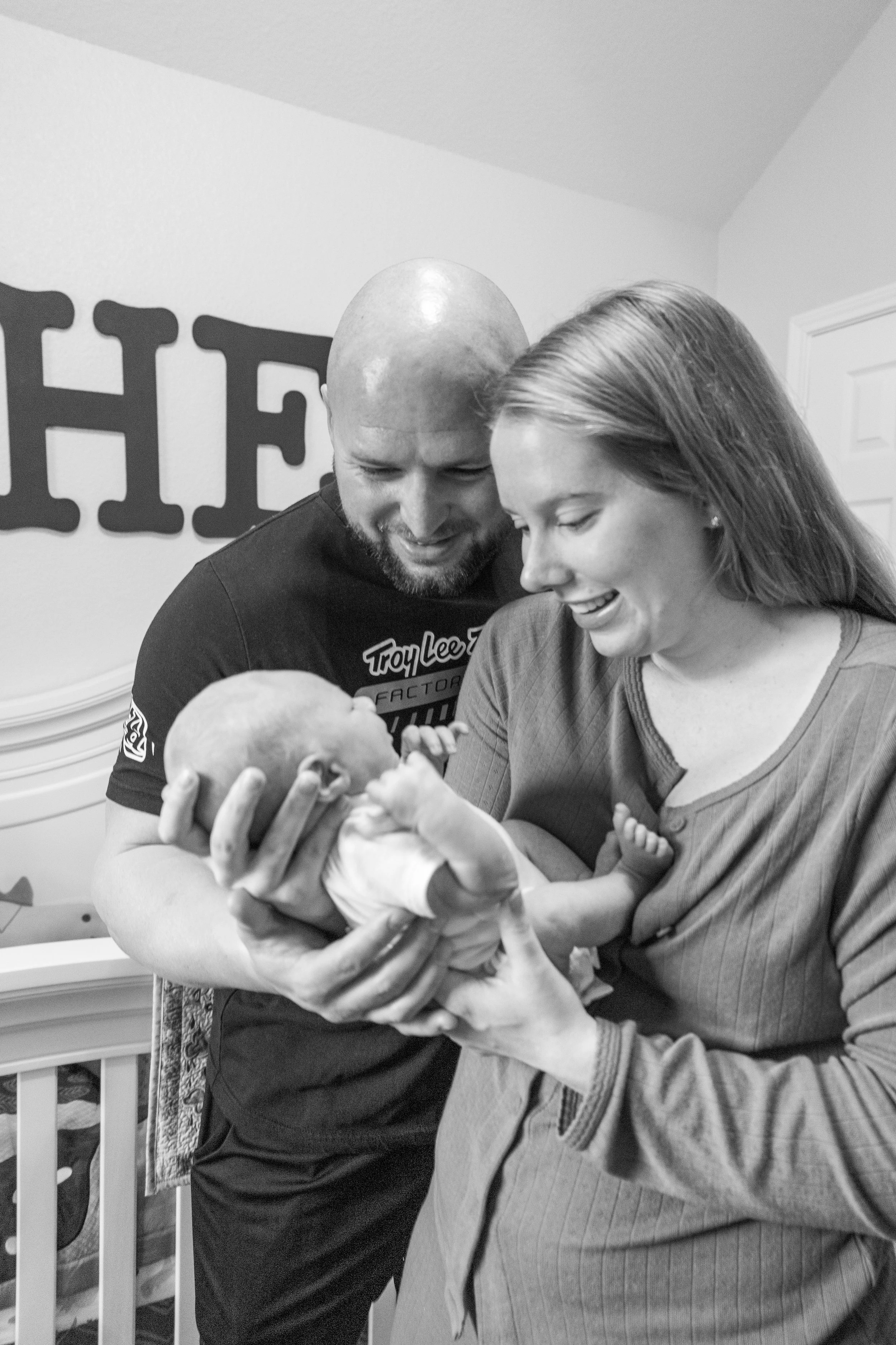 A couple holding a newborn baby, looking at the baby with smiles. The setting appears to be a nursery, with a crib and family decor in the background.