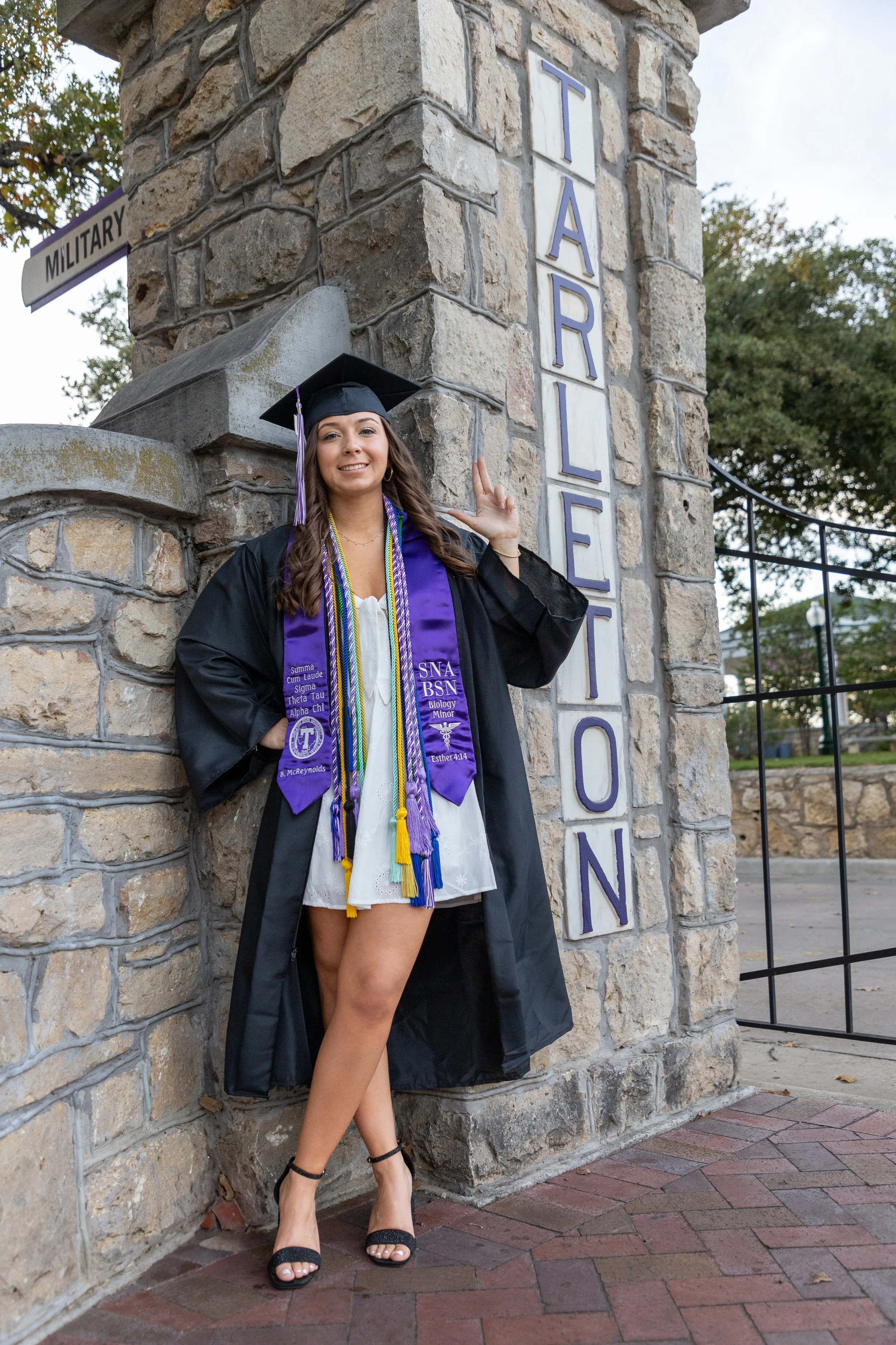A young woman in a graduation cap and gown standing next to a stone pillar with a "TARLETON" sign. She is smiling, wearing a purple stole and multiple honor cords, and is posed with one finger pointing up.
