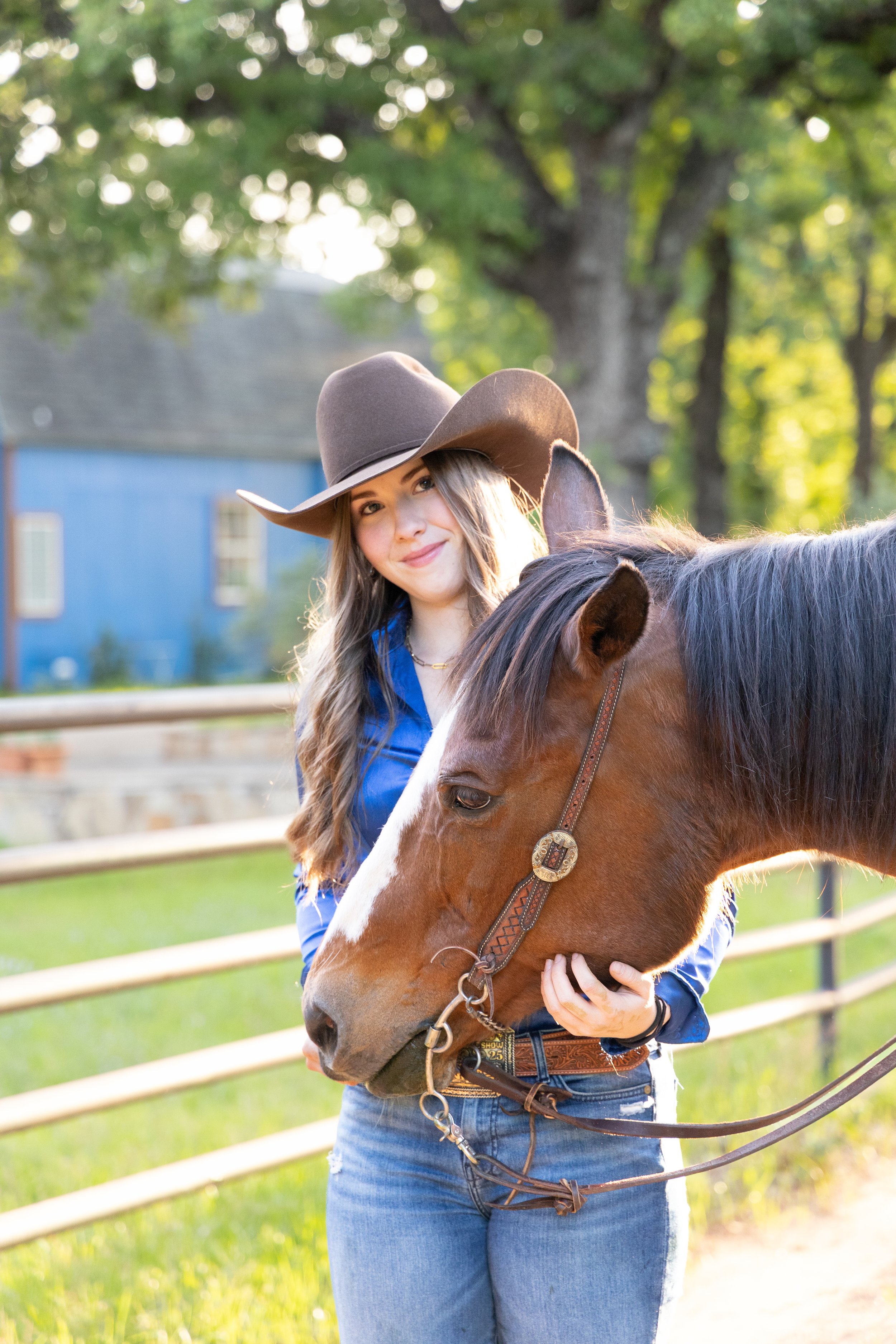 A young woman with long light brown hair, wearing a brown cowboy hat, a blue denim shirt, and jeans, is standing outdoors with a brown horse. She is smiling and holding the horse's head gently, with a background of a green field, trees, and a blue bu