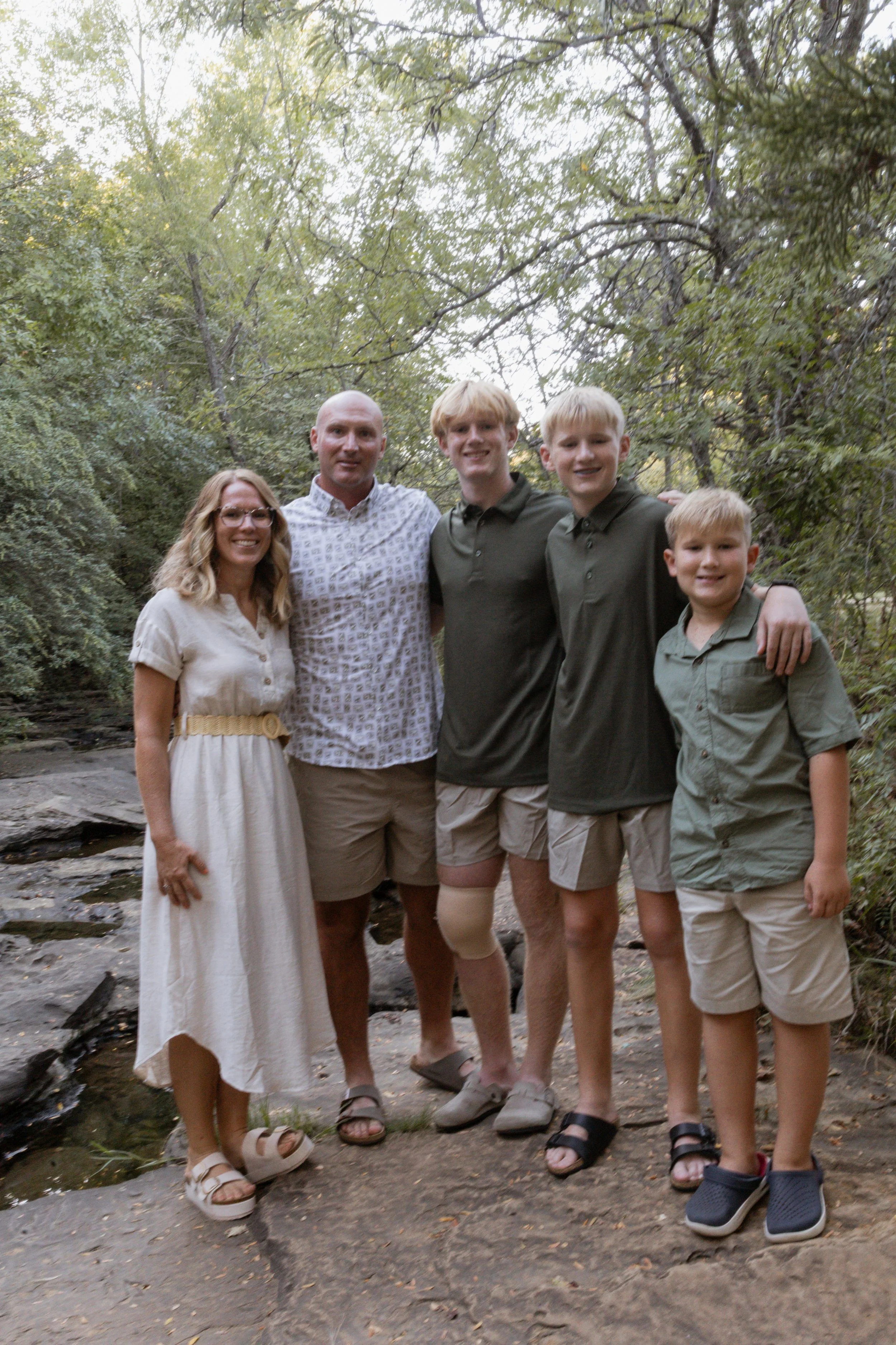 Family of five outside in a wooded area, standing on rocks near a stream, smiling at the camera.