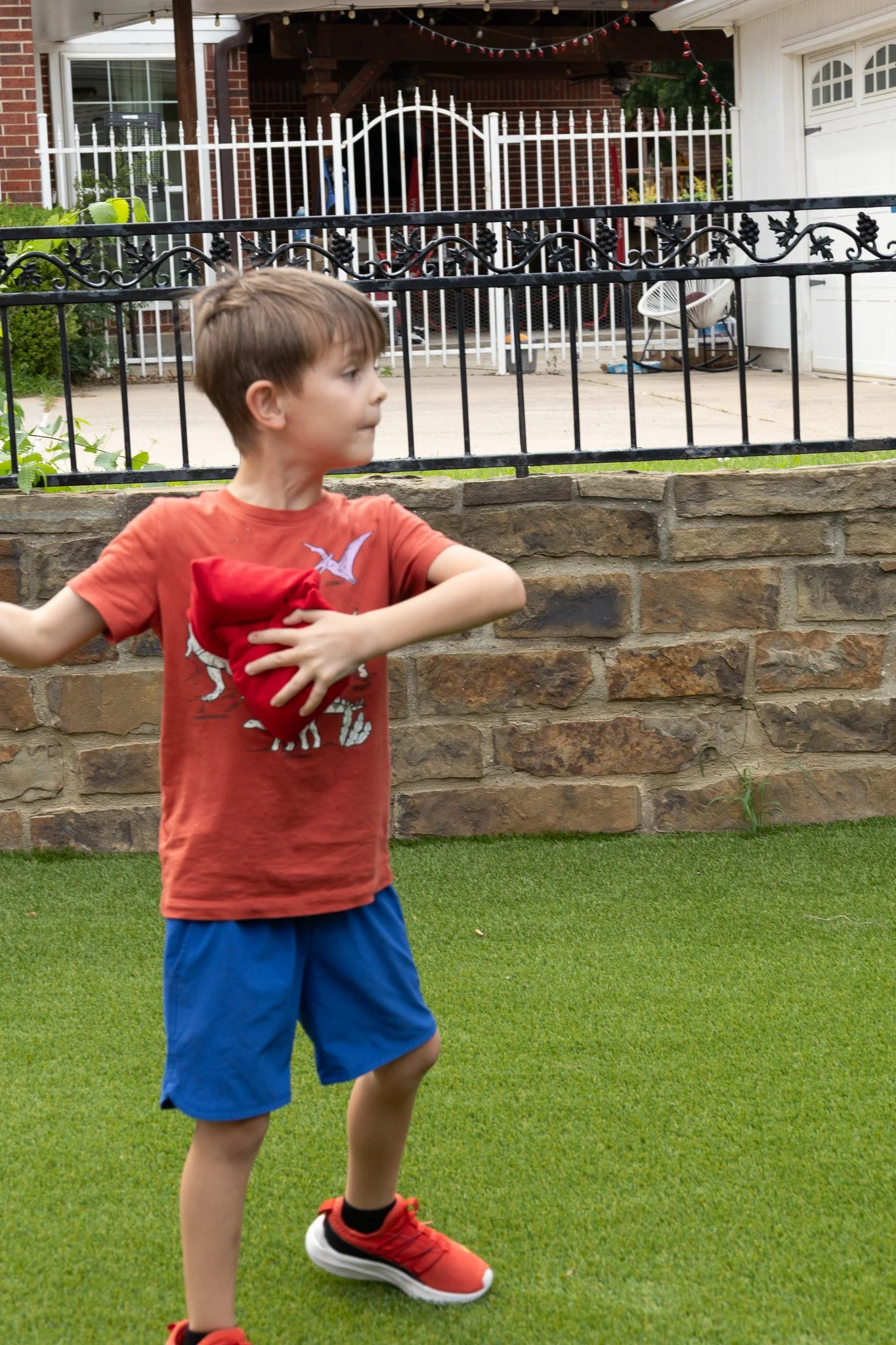 A young boy wearing an orange T-shirt, blue shorts, and red sneakers stands on a green artificial grass lawn, holding a red hat against his chest. There is a stone wall and a black metal fence behind him, with a residential backyard containing patio 