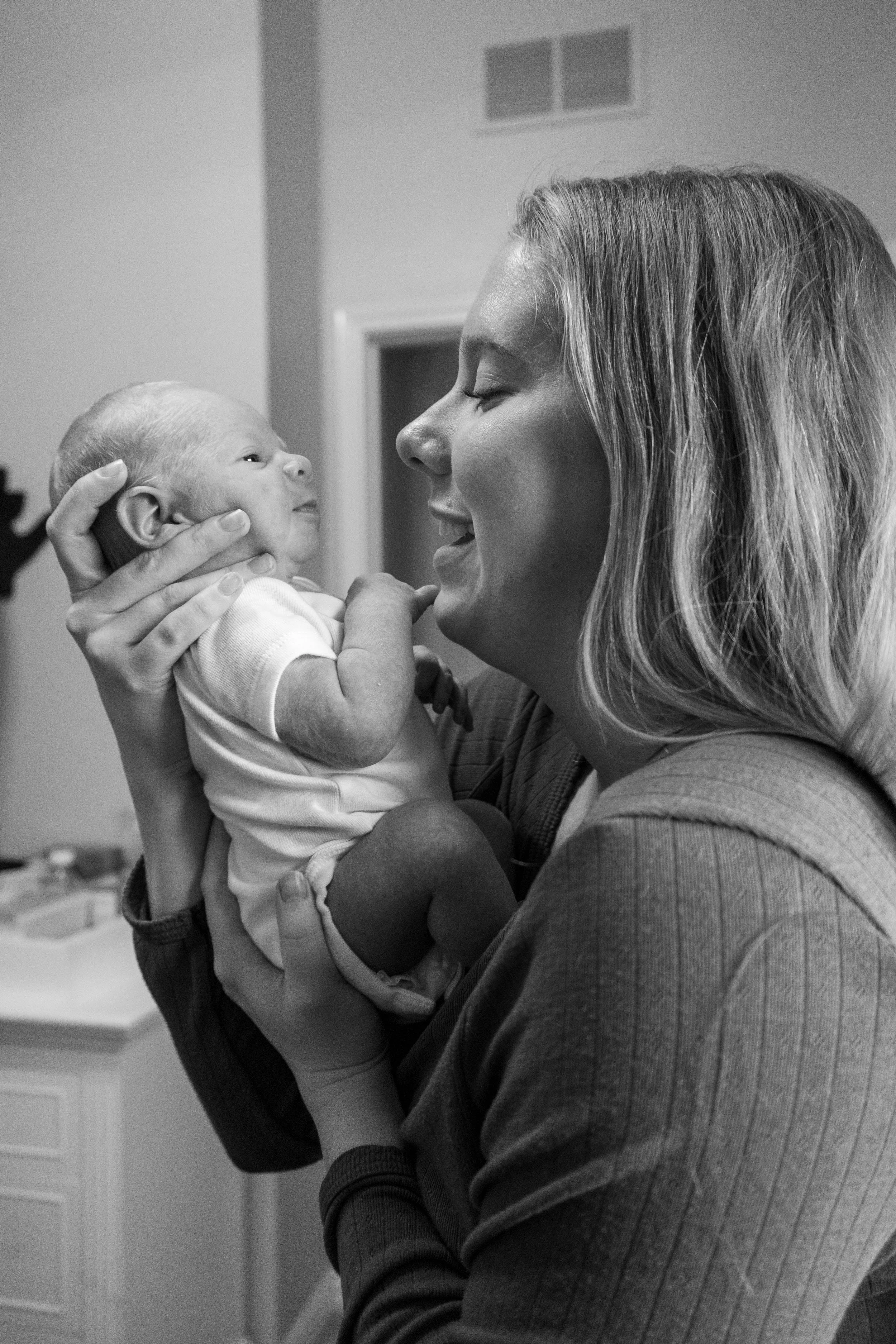 A woman holding a baby close, both smiling and looking at each other in a warmly lit room.