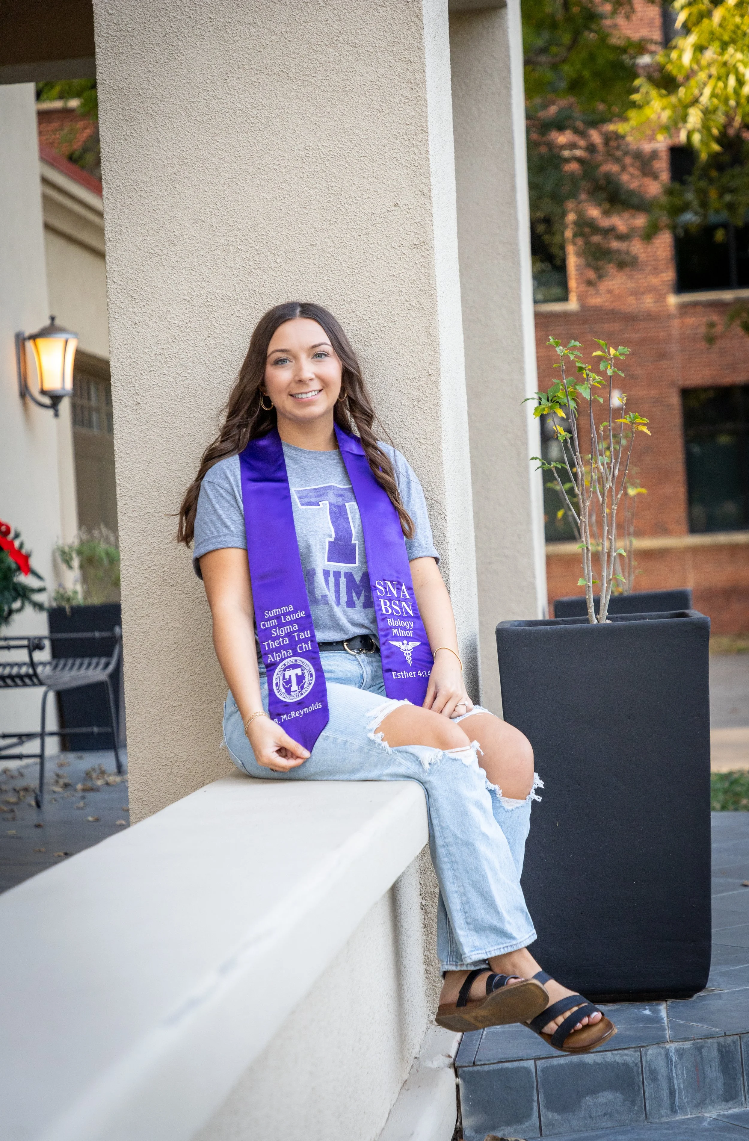 A young woman sitting on a ledge outdoors, wearing a grey T-shirt and ripped jeans, with a purple sorority stole and smiling at the camera.