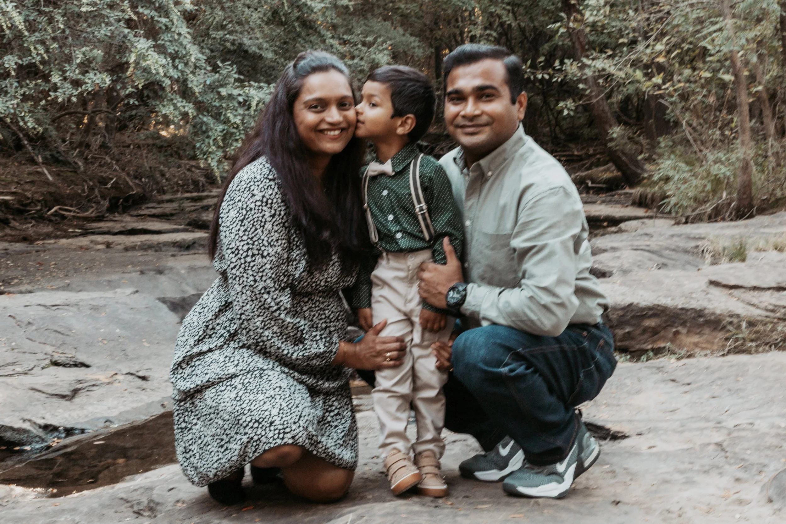 A family of three, a woman, a man, and a young boy, outdoors on a rocky area with trees in the background. The woman is kneeling, smiling, wearing a patterned dress. The boy in the middle is kissing her cheek, wearing a green shirt with suspenders an