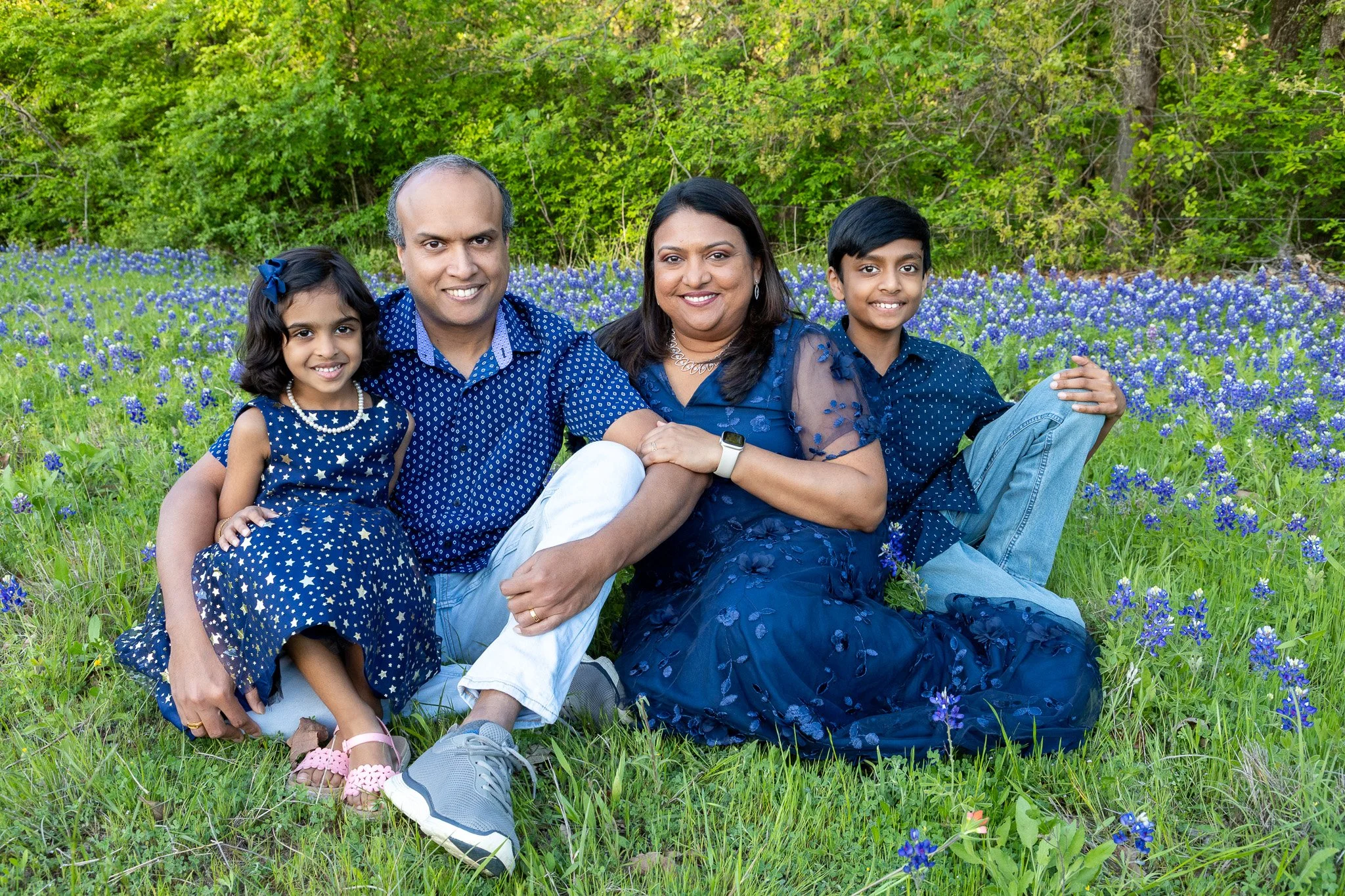 Family of four sitting on grass in a field of blue flowers with green trees in the background. The family includes a young girl, a man, a woman, and a boy, all smiling and dressed in blue.