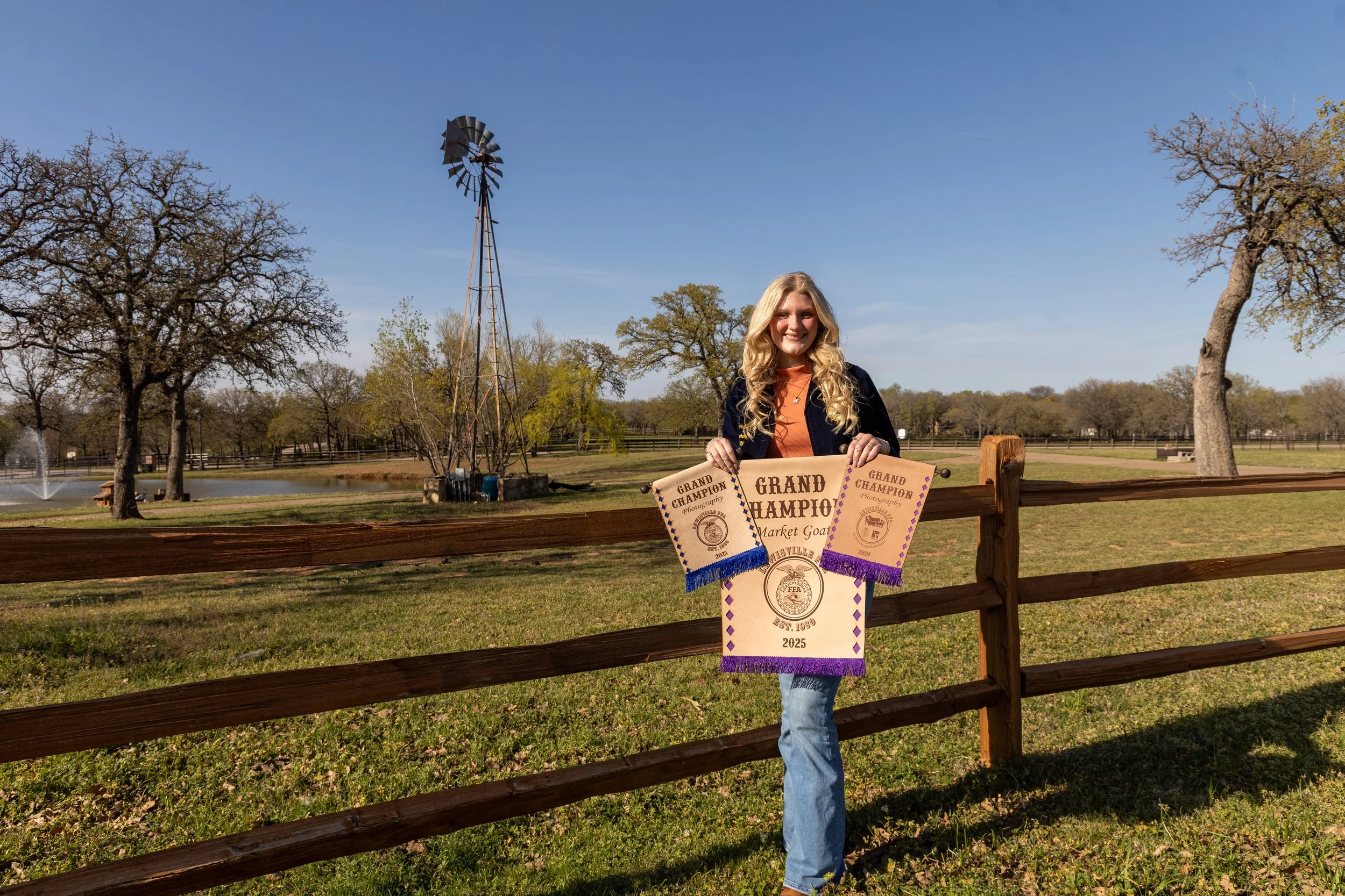A woman standing outdoors near a wooden fence holding a large bike riding champion banner with purple fringe, a smaller banner with blue fringe, and a silver medal in the center. The background features a park with trees, a fountain, and a windmill u
