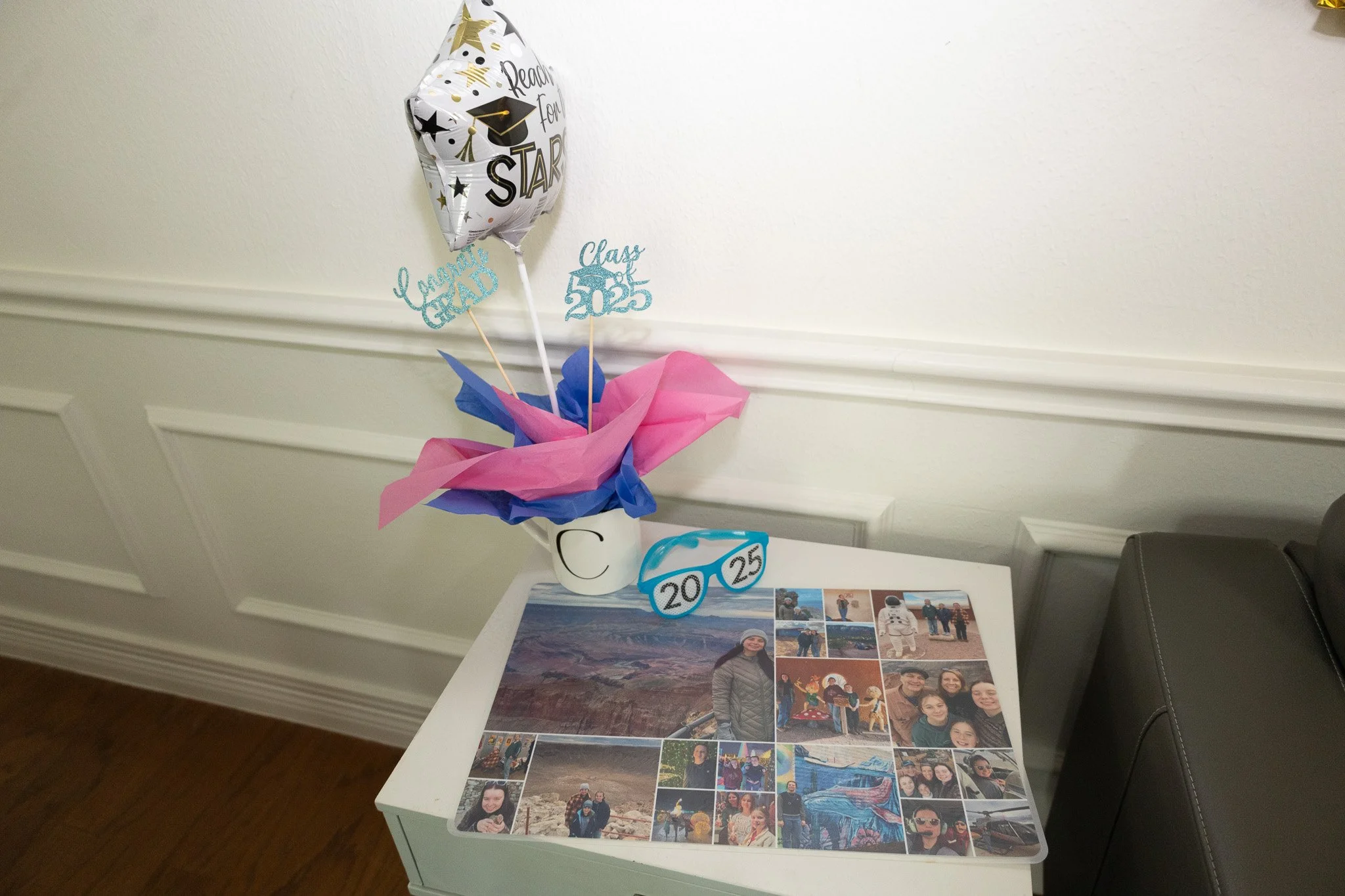 Decorative graduation-themed arrangement with a balloon, paper flowers, and signs reading "Class of 2025" and "Graduate Grad." The arrangement is in a white cup on a small table with a collage of photos displayed on it.