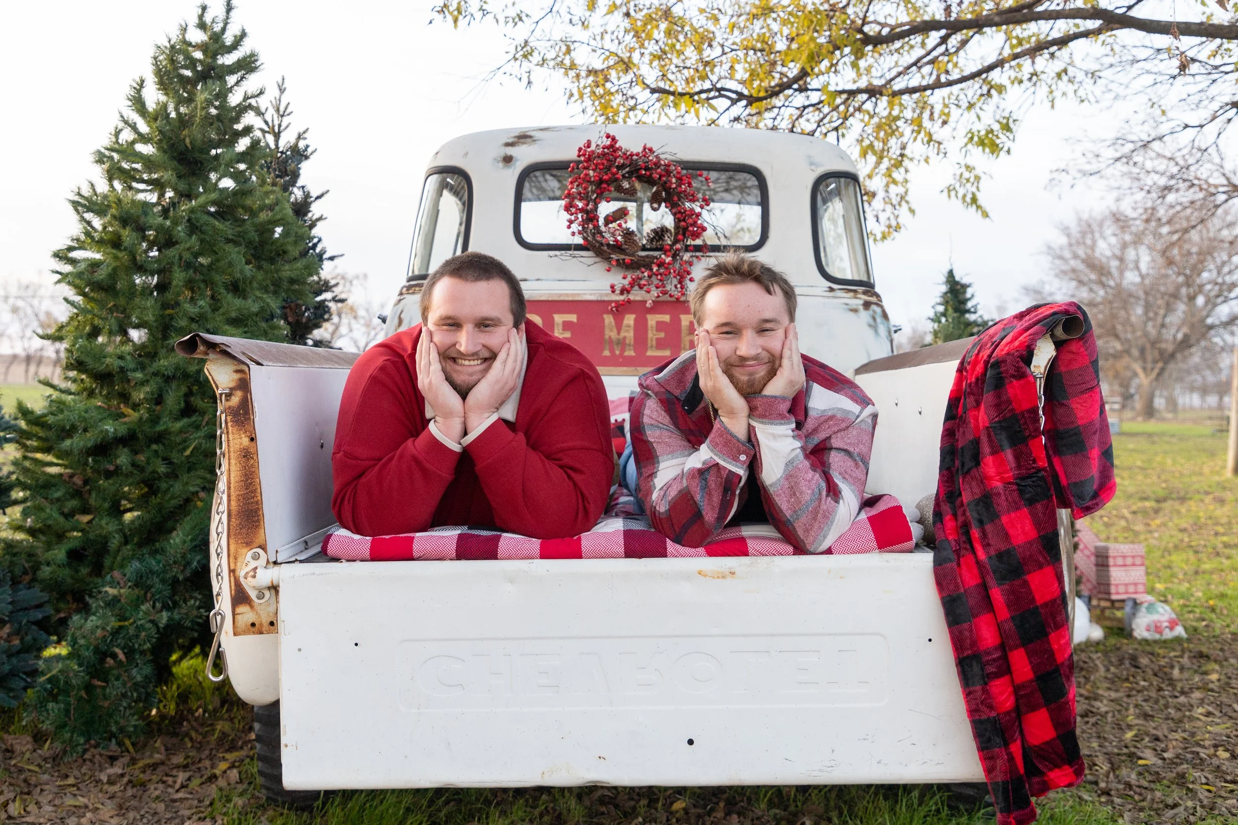 Two smiling men lying in the bed of a vintage white truck decorated with a Christmas wreath, with pine trees and wrapped presents nearby, during the holiday season.