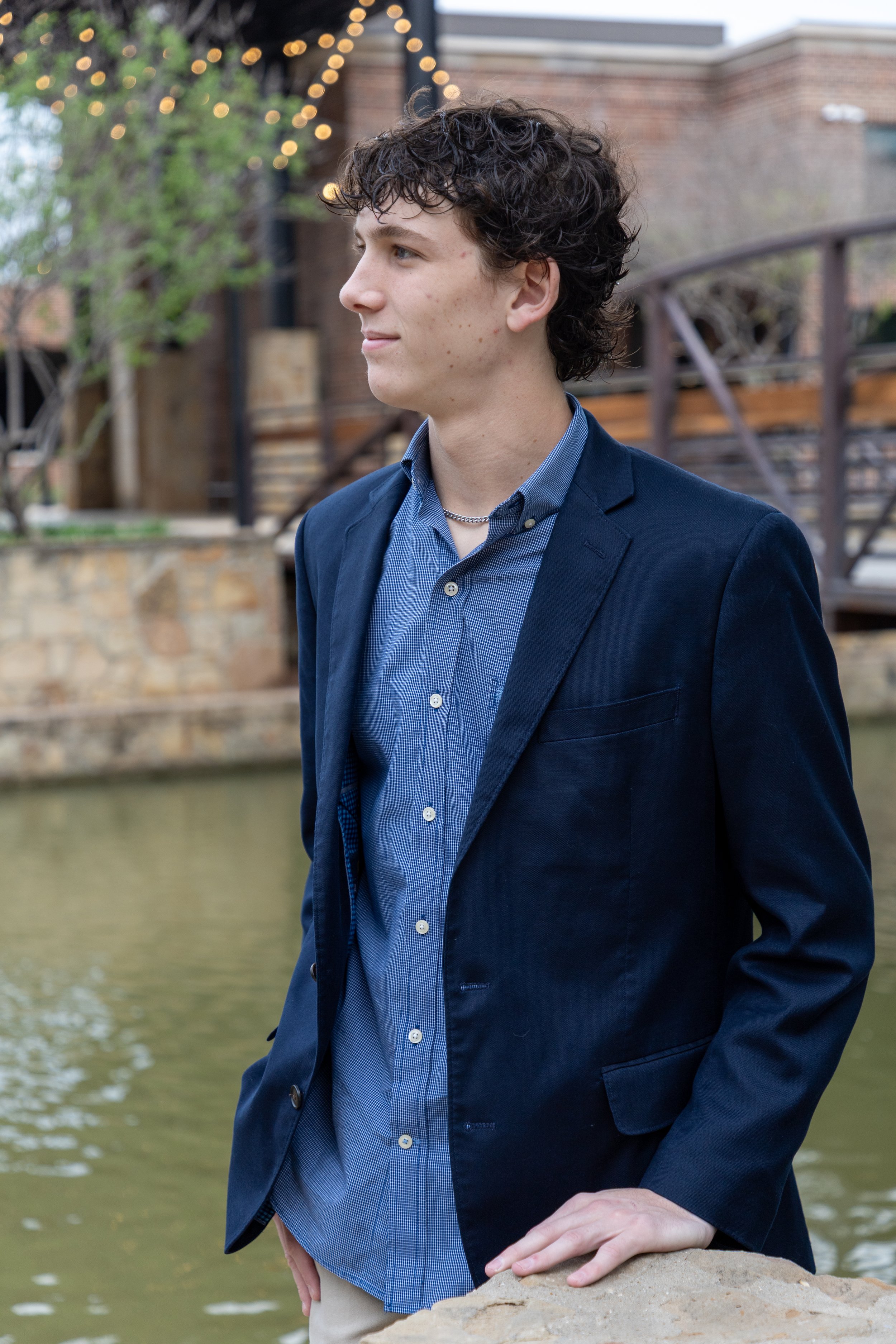 A young man with curly brown hair, wearing a navy blazer and blue shirt, standing by a body of water with a stone wall and building with string lights in the background.