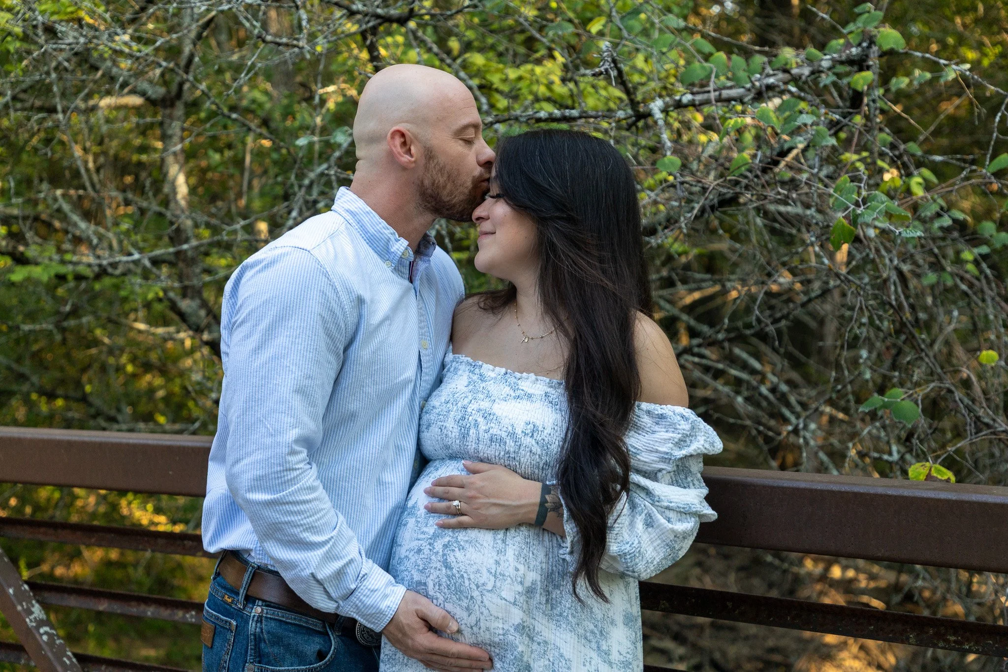 A pregnant woman and a man kissing on the forehead in an outdoor wooded area, with the woman touching her belly.