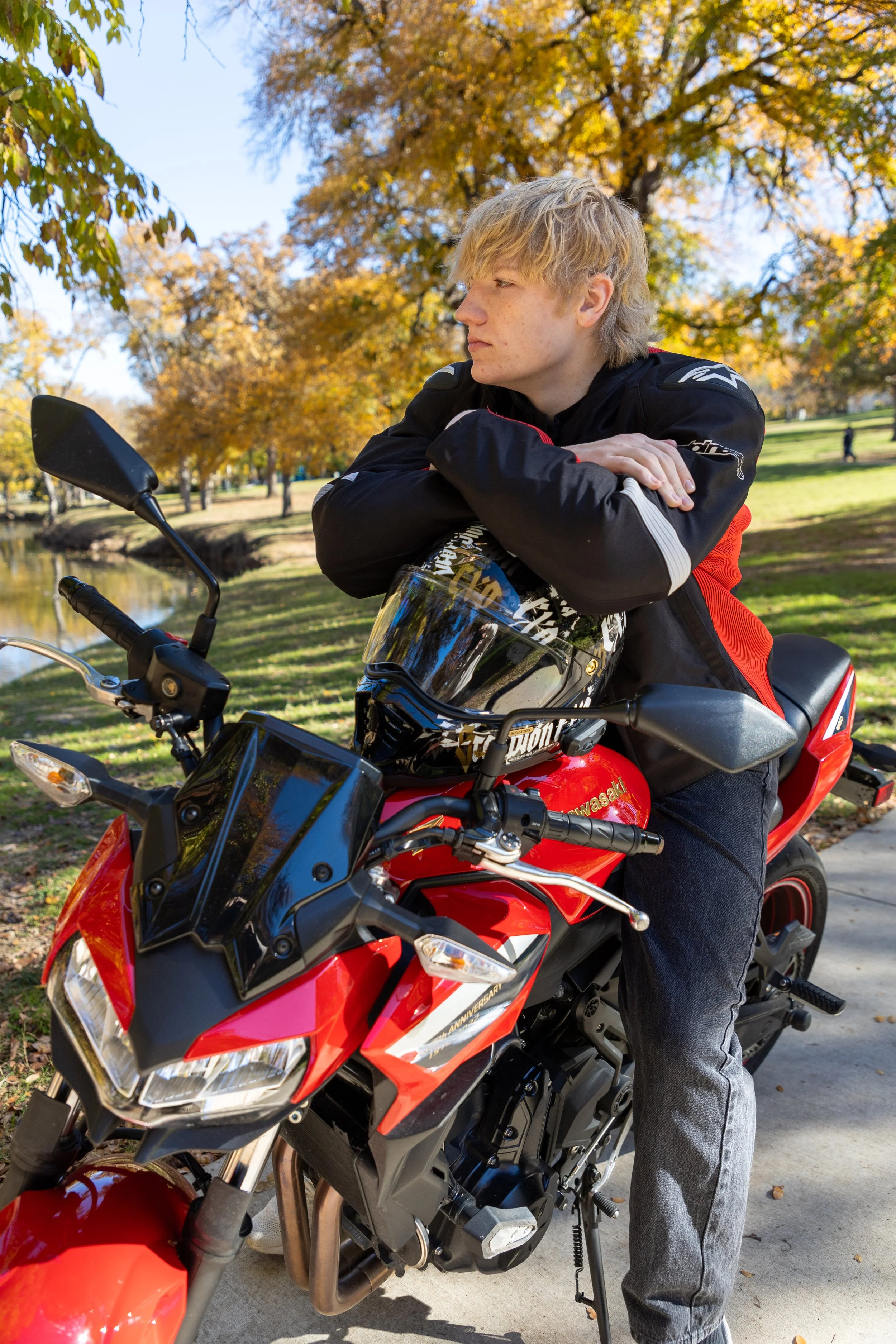 Young man with blonde hair sitting cross-armed on a red Kawasaki motorcycle, looking to the side, in a park with autumn trees and a lake.