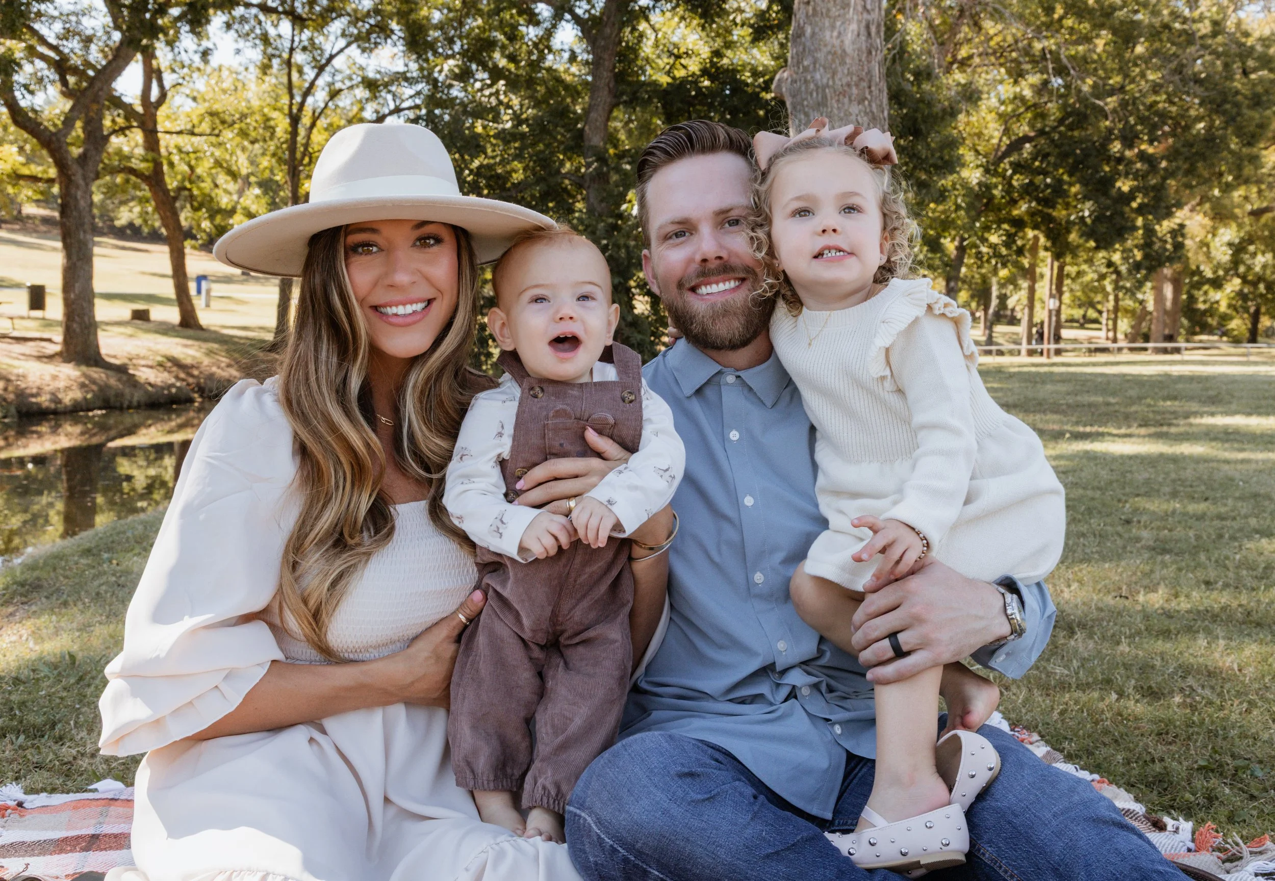 Family of four sitting outdoors on a blanket, smiling, near a lake and trees in a park.
