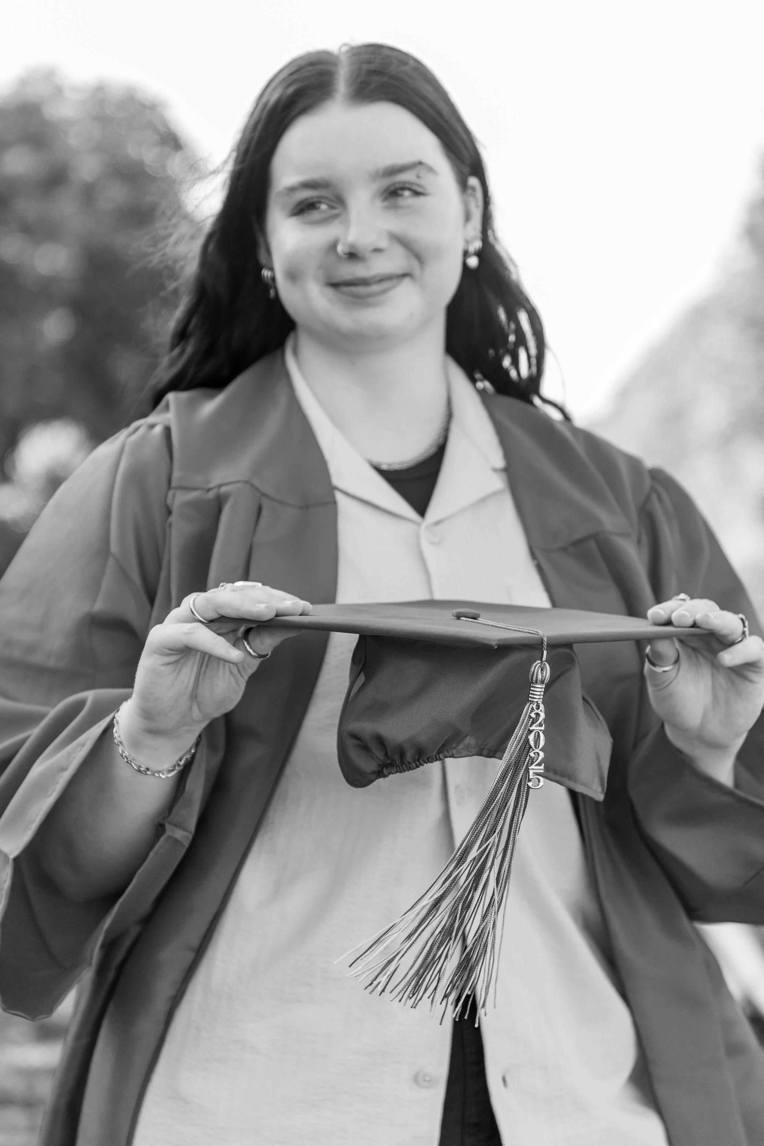 A young woman in graduation attire holding a graduation cap with a tassel marked 2025, smiling outdoors.