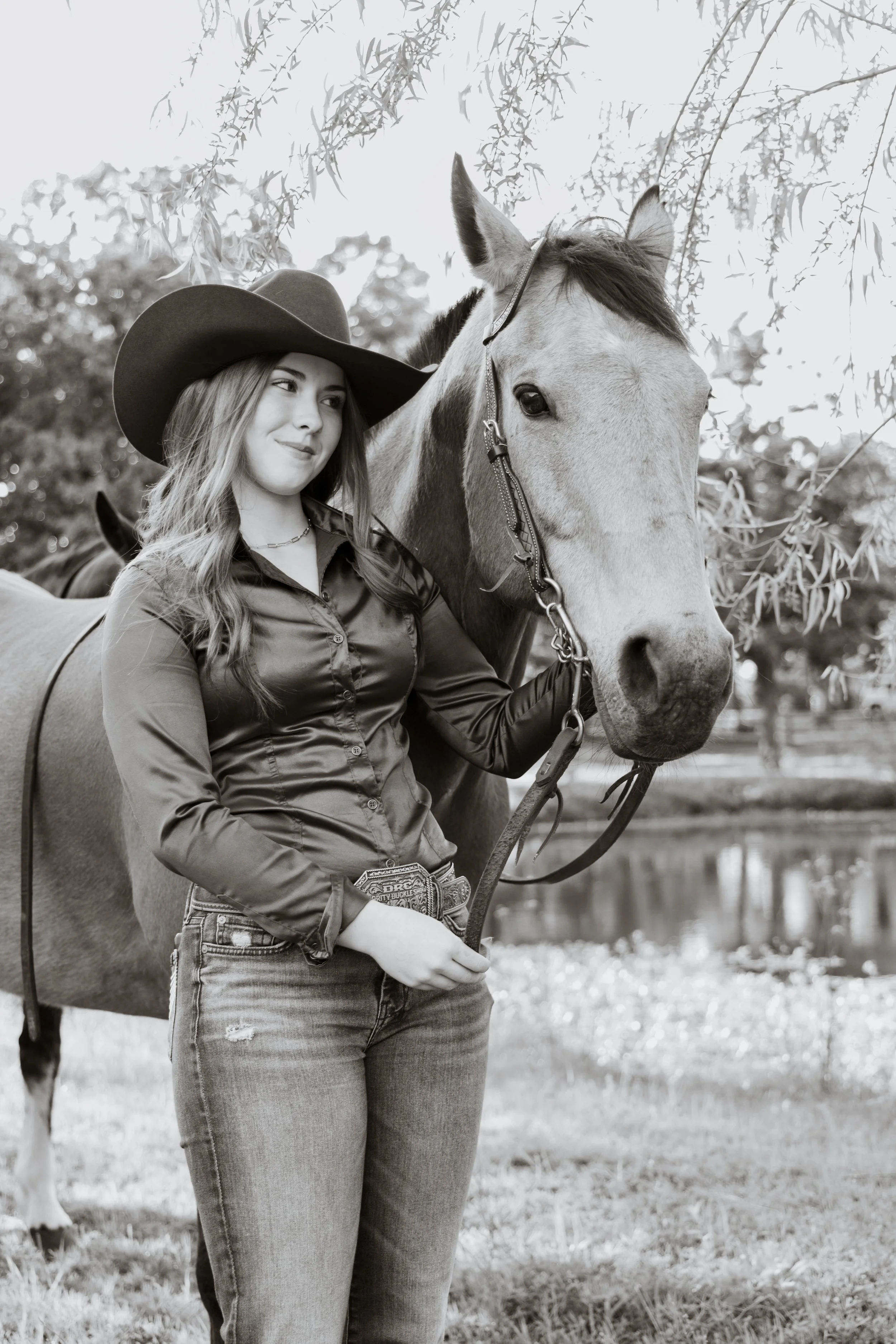 A young woman with long hair wearing a cowboy hat, plaid shirt, and jeans, standing next to a horse, holding its reins outdoors by a pond, with trees and leaves overhead in black and white.
