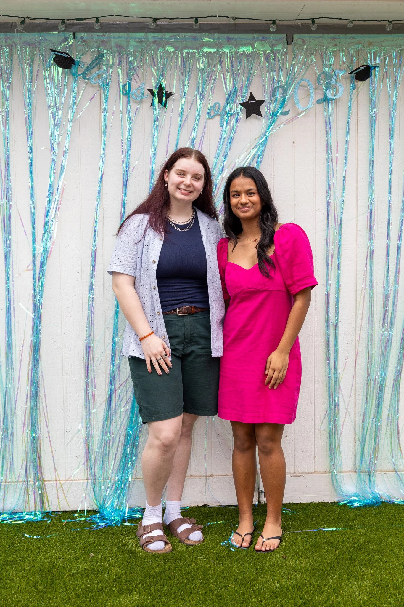 Two women standing together in front of a decorative backdrop with metallic streamers and black star cutouts. One woman has long red hair and is wearing a navy top, shorts, and sandals. The other woman has long dark hair and is wearing a bright pink 