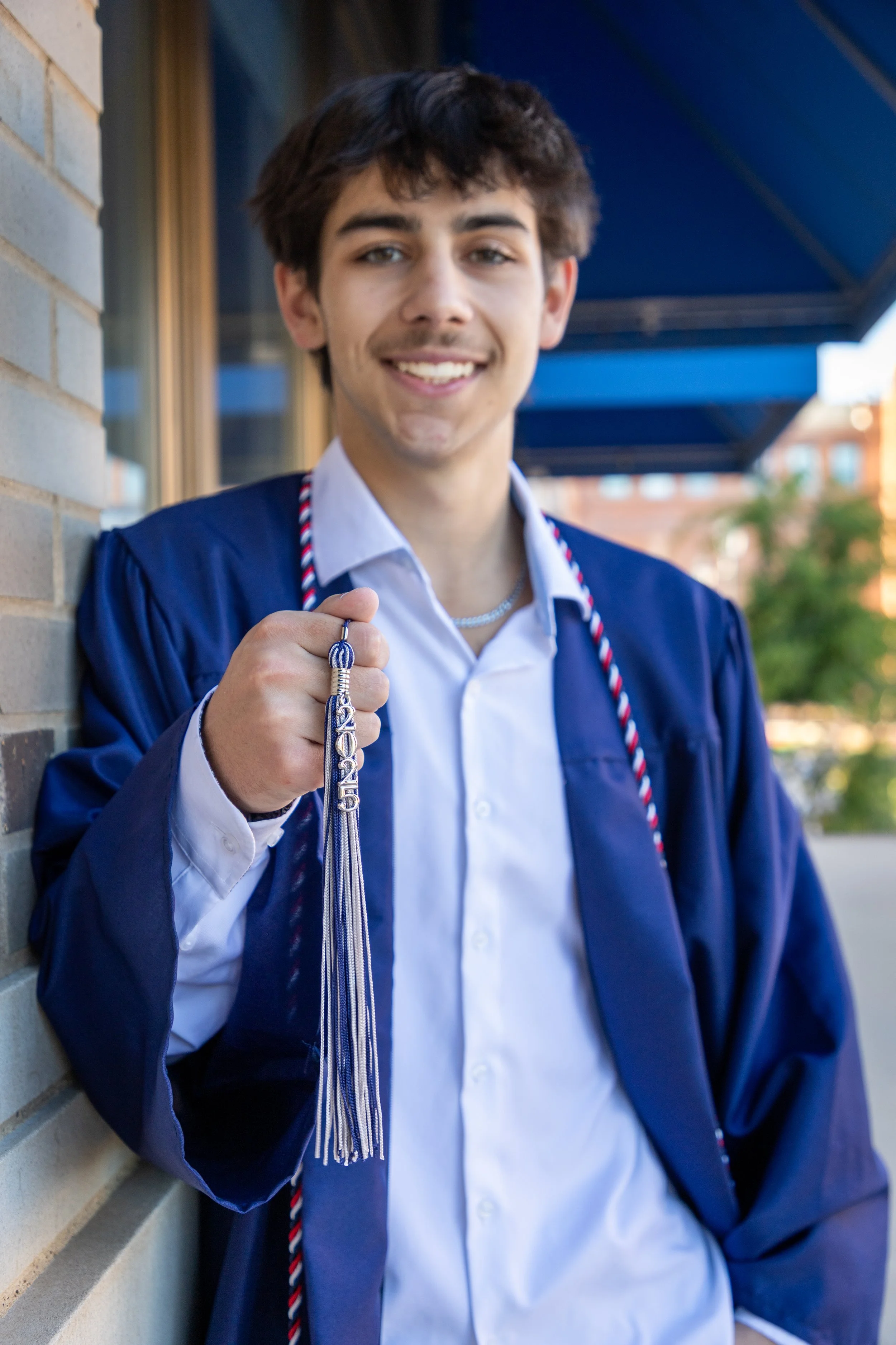 Young man in a graduation gown holding a tassel, smiling outdoors.