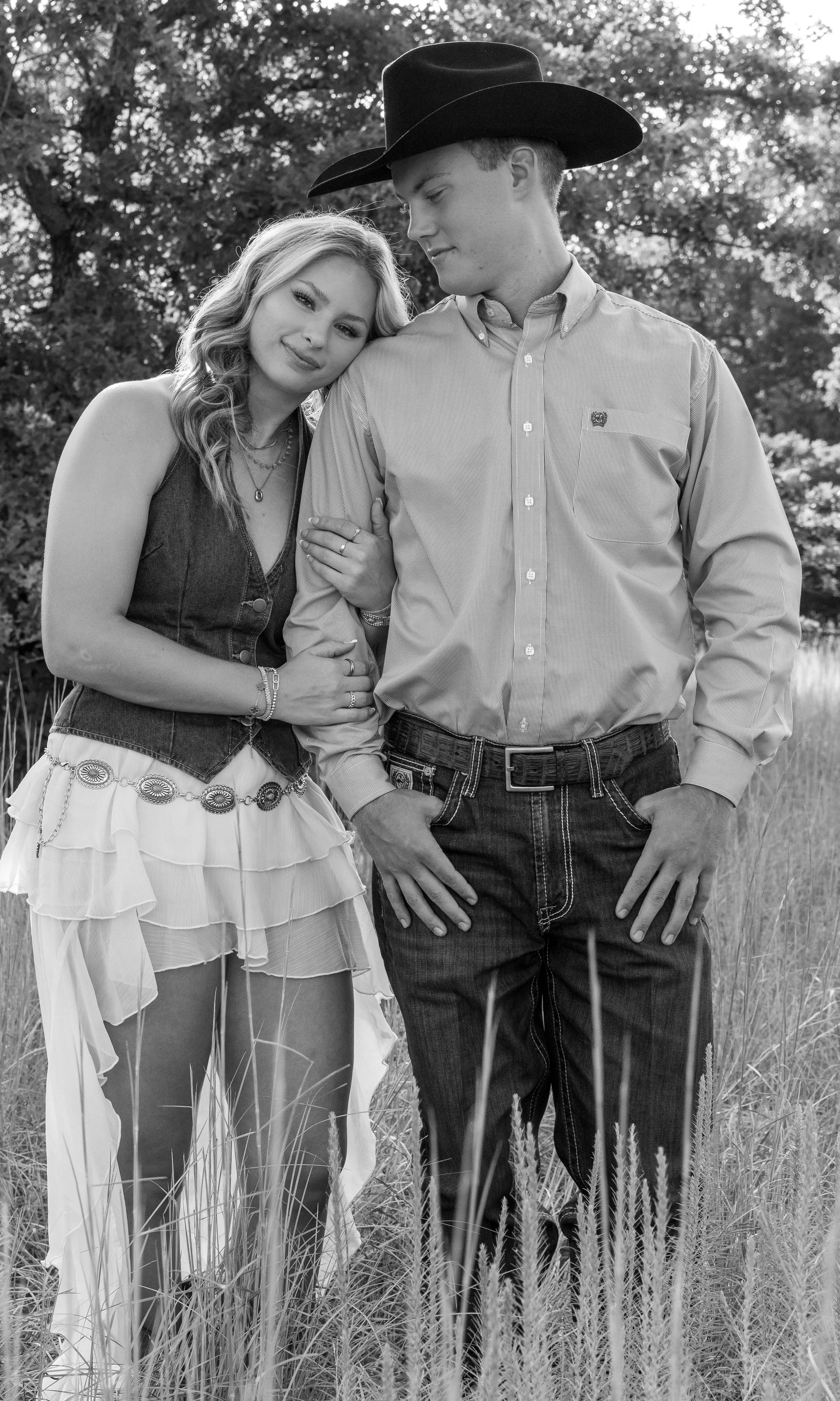 Black and white photo of a couple in a field of tall grass. The woman has long wavy hair, wears a sleeveless vest and a flowing skirt with decorative chain details, and is resting her head on the man's shoulder. The man wears a button-up shirt, jeans