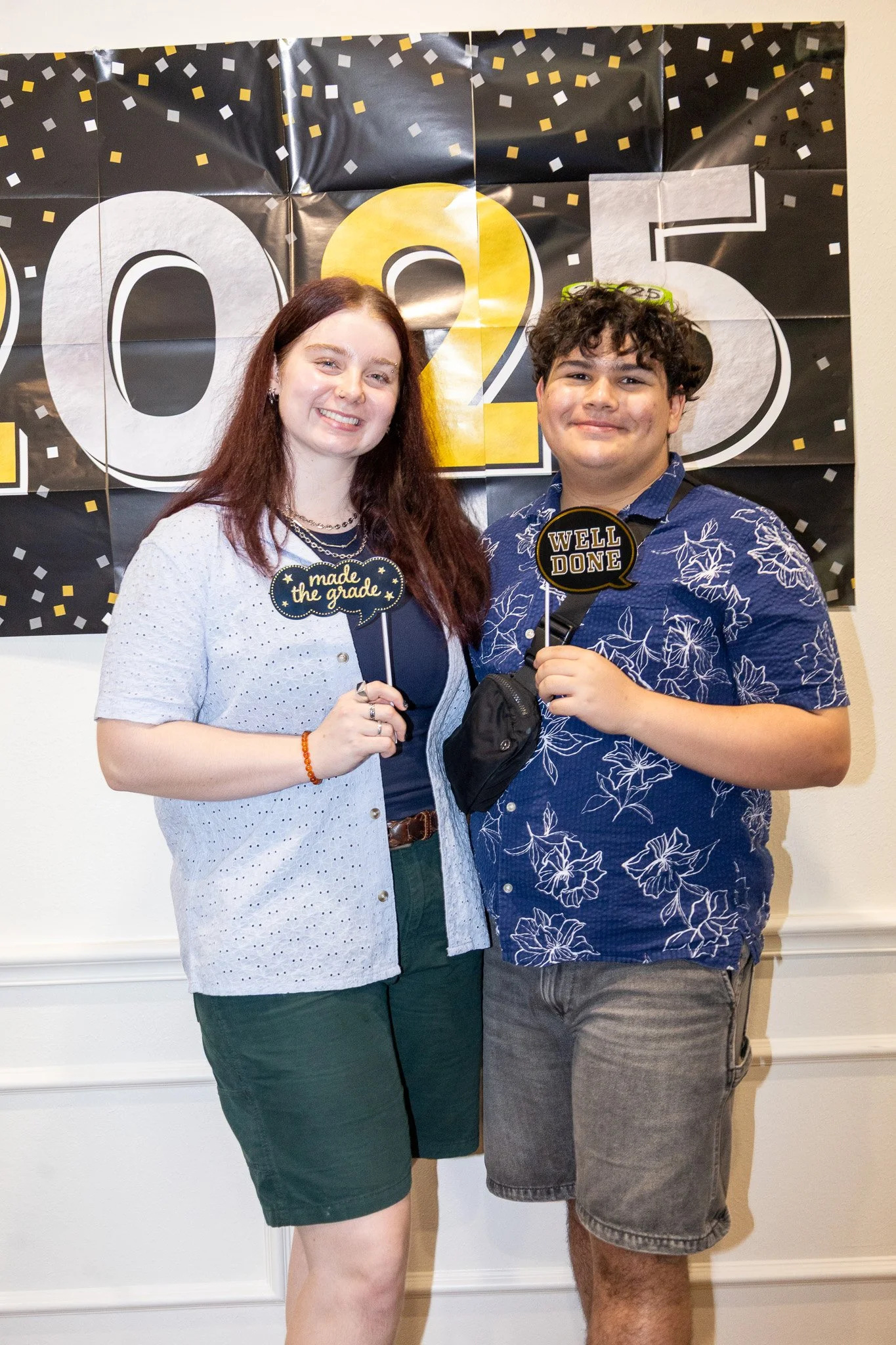 Two young adults smiling at a graduation celebration, holding signs that say 'Made the grade' and 'Well Done', standing in front of a black and gold '2023' banner with confetti design.