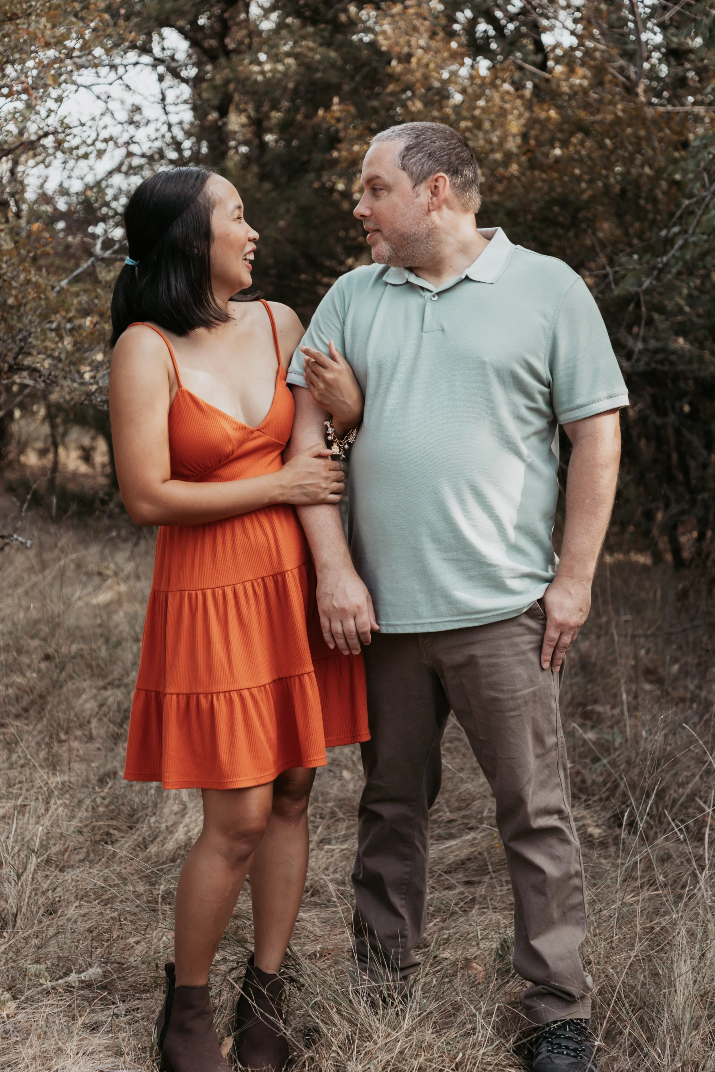 A couple sharing a joyful moment outdoors, standing on dry grass with trees in the background. The woman wears an orange dress and the man wears a light green polo shirt and brown pants. They are looking at each other affectionately.