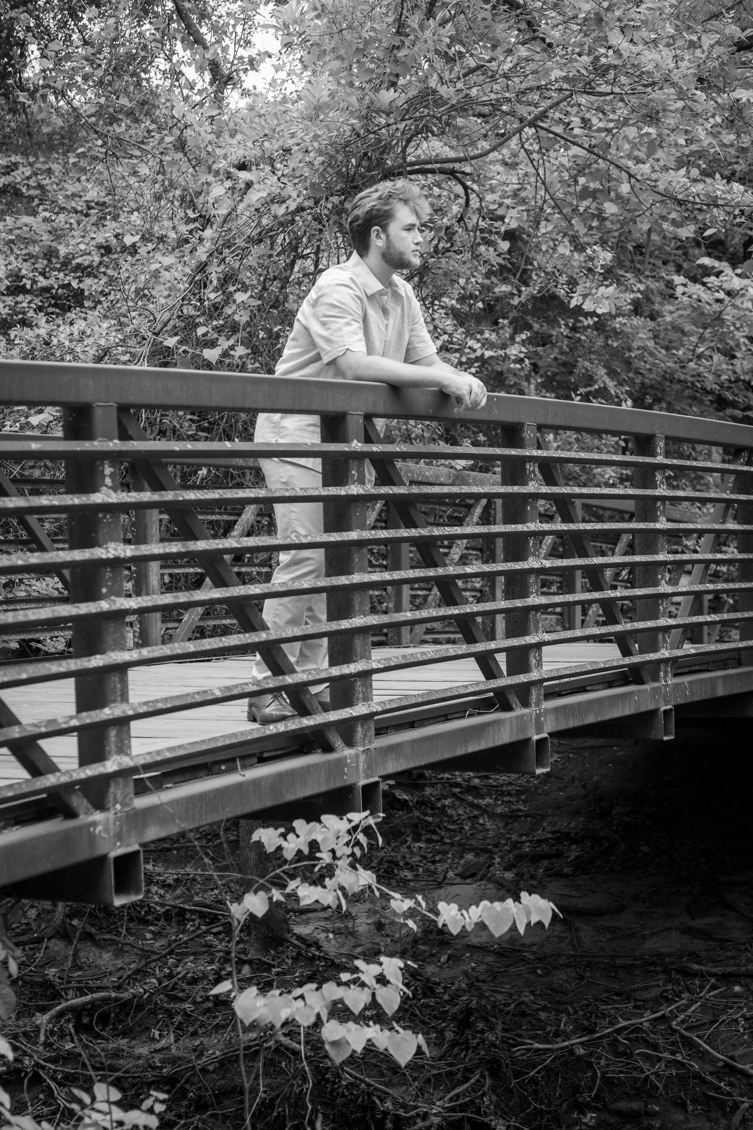A man with styled hair and a beard is leaning on a metal railing of a bridge, looking into the distance. The bridge is in a wooded area with trees and leaves in the background. The photo is in black and white.