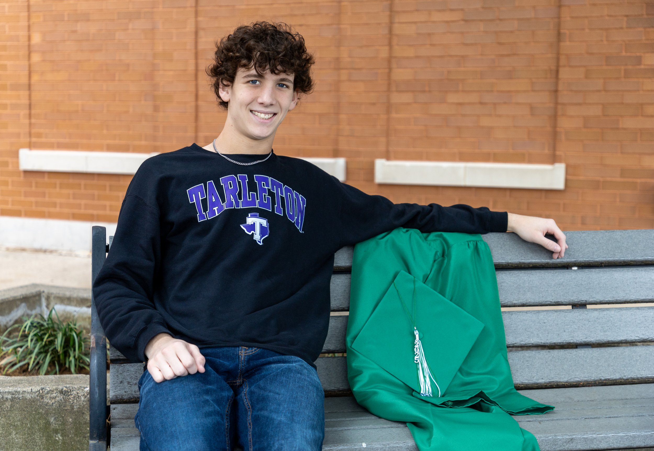 A young man with curly hair wearing a black T-shirt with 'Tarleton' written on it and a blue logo, sitting on a gray park bench with a folded green graduation gown and cap beside him, outside in front of a brick building.