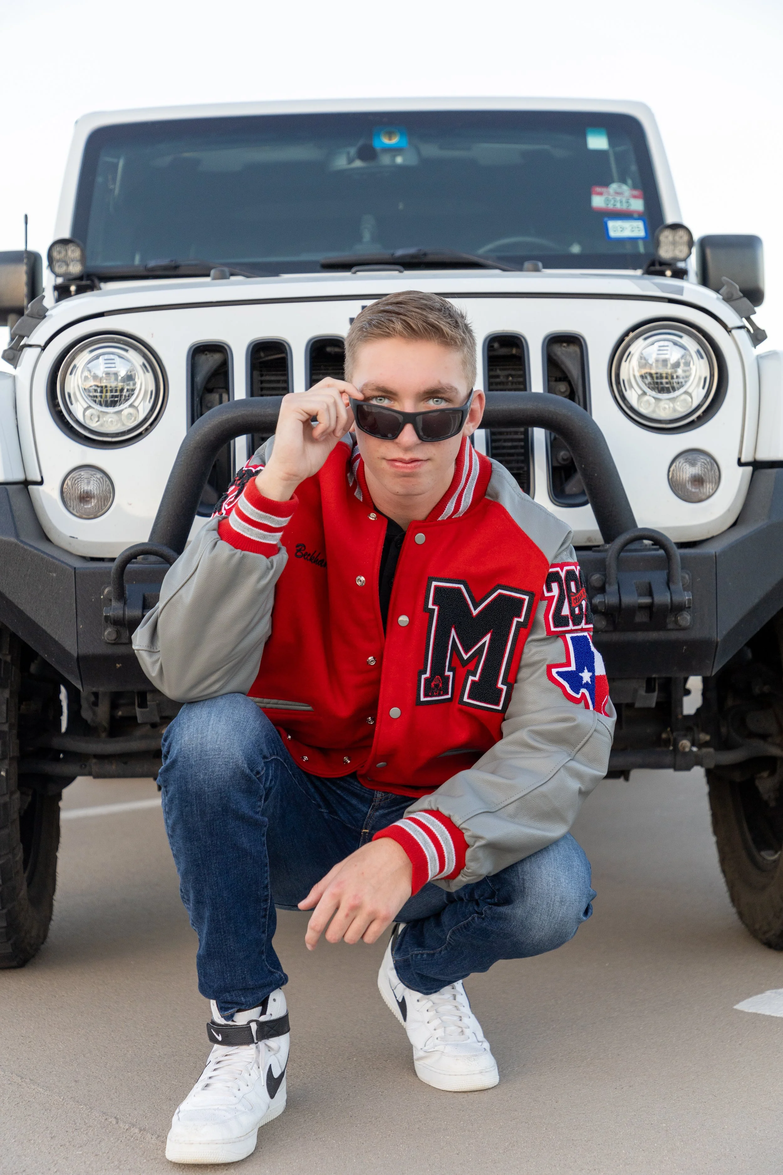 A young man crouching in front of a white Jeep, wearing a red and gray varsity jacket, blue jeans, white Nike sneakers, and sunglasses, looking at the camera.