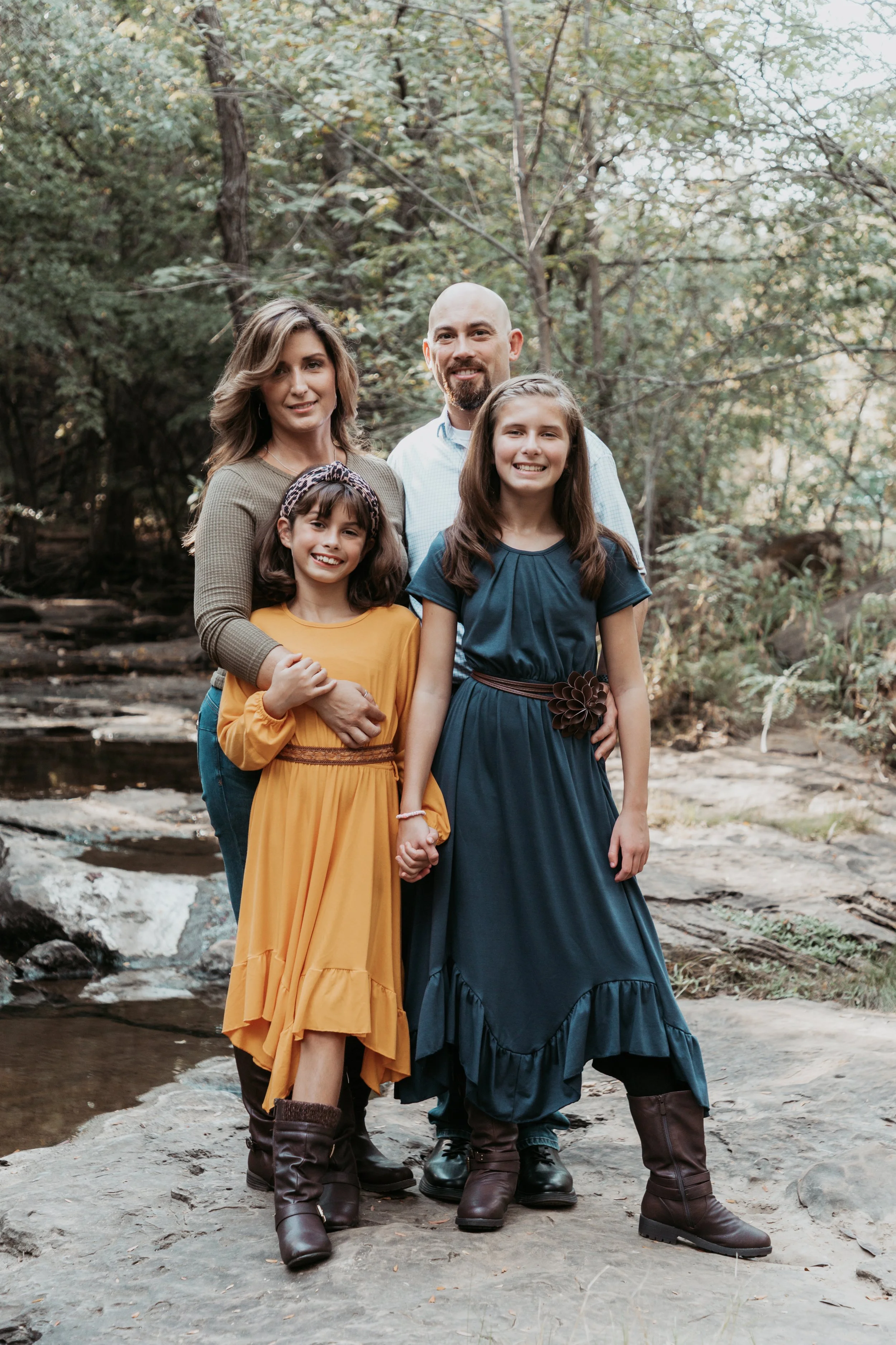A family of four standing outdoors in a wooded area, with two young girls in front and a man and woman behind them, all smiling and holding hands.