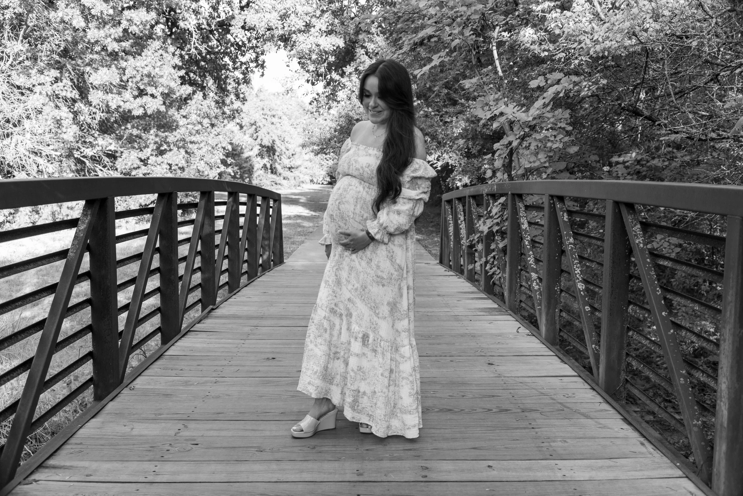 A pregnant woman standing on a wooden bridge surrounded by trees.