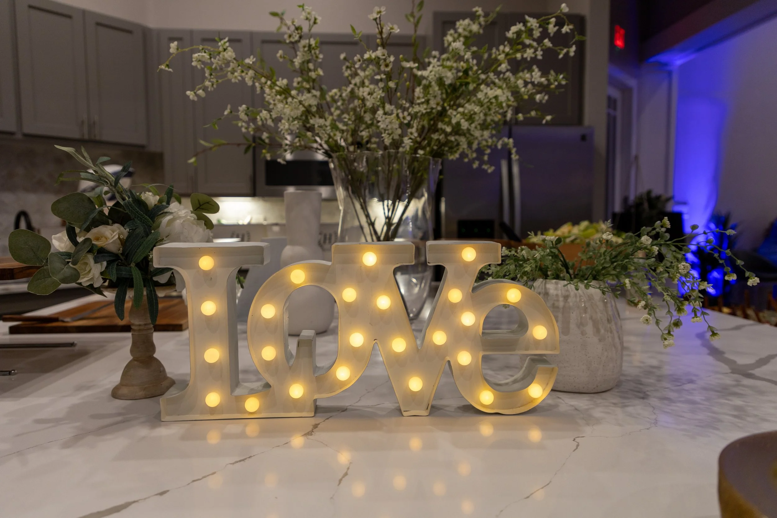 Decorative illuminated sign spelling 'LOVE' on a marble table with floral arrangements in a modern kitchen or dining area