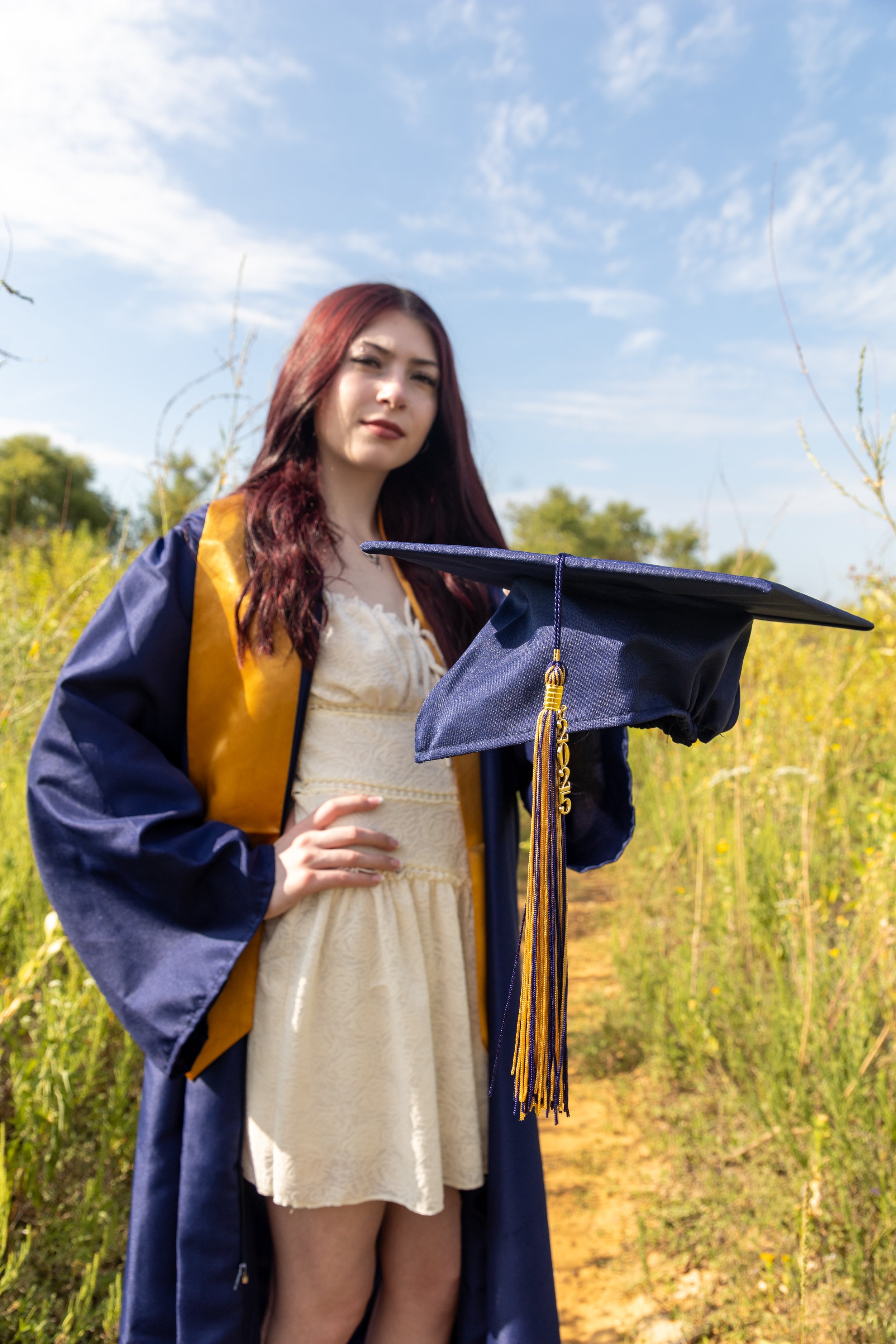 A young woman in a graduation gown holding a cap with a yellow tassel, standing outdoors on a sunny day with a clear blue sky and green trees in the background.