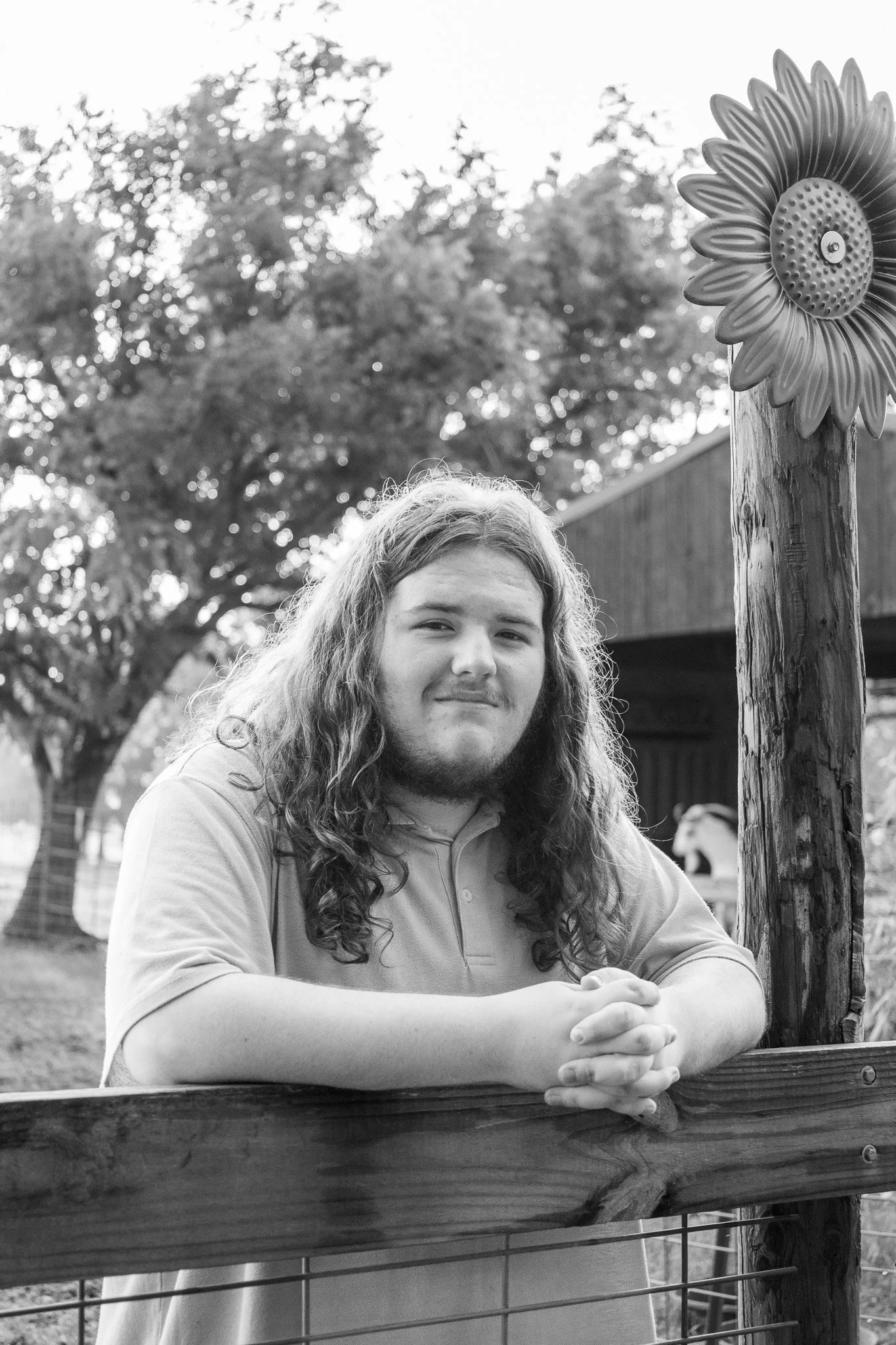 A young man with long, wavy hair and a beard leaning on a wooden fence outdoors, with a tree and a small shed in the background, in black and white.