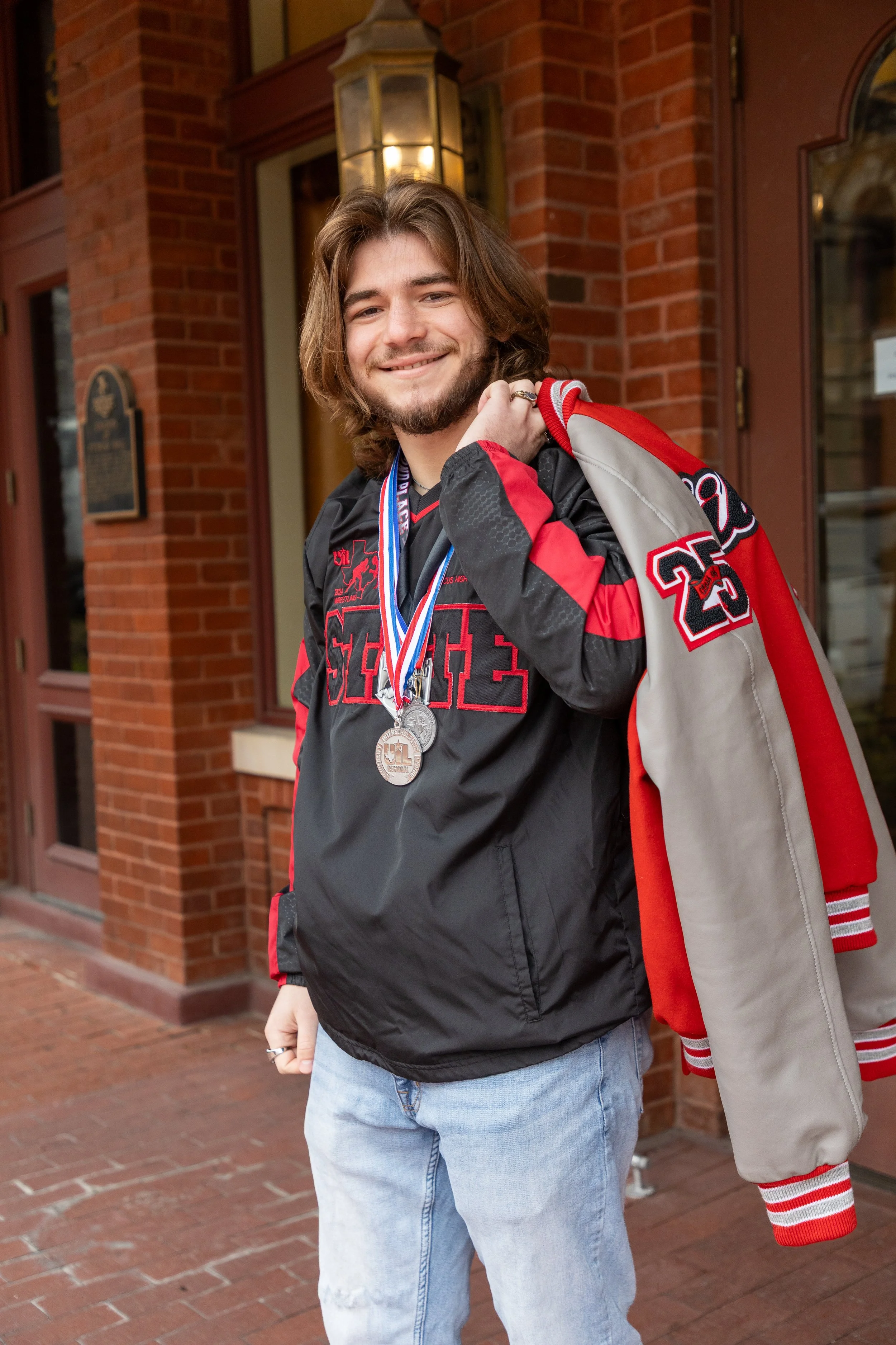 A young man with long brown hair and a beard smiling, wearing a black and red sports jacket with the letter 'S' on it, two medals around his neck, holding a red and gray school jacket over his shoulder, standing outside a school building made of red 