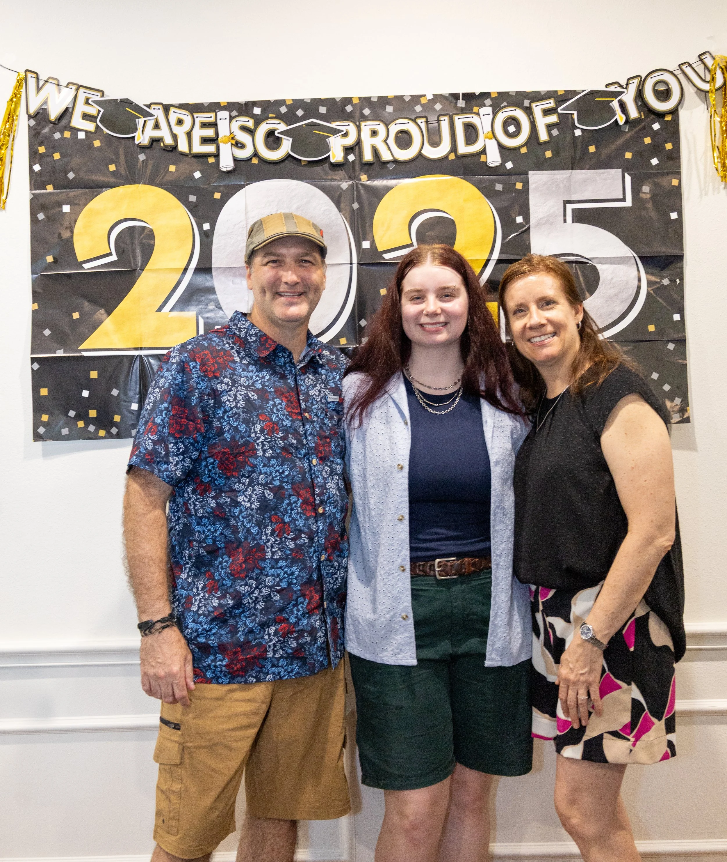 Three people celebrating a graduation, standing in front of a black and gold banner that reads 'We are so proud of you 2025', with confetti decoration.