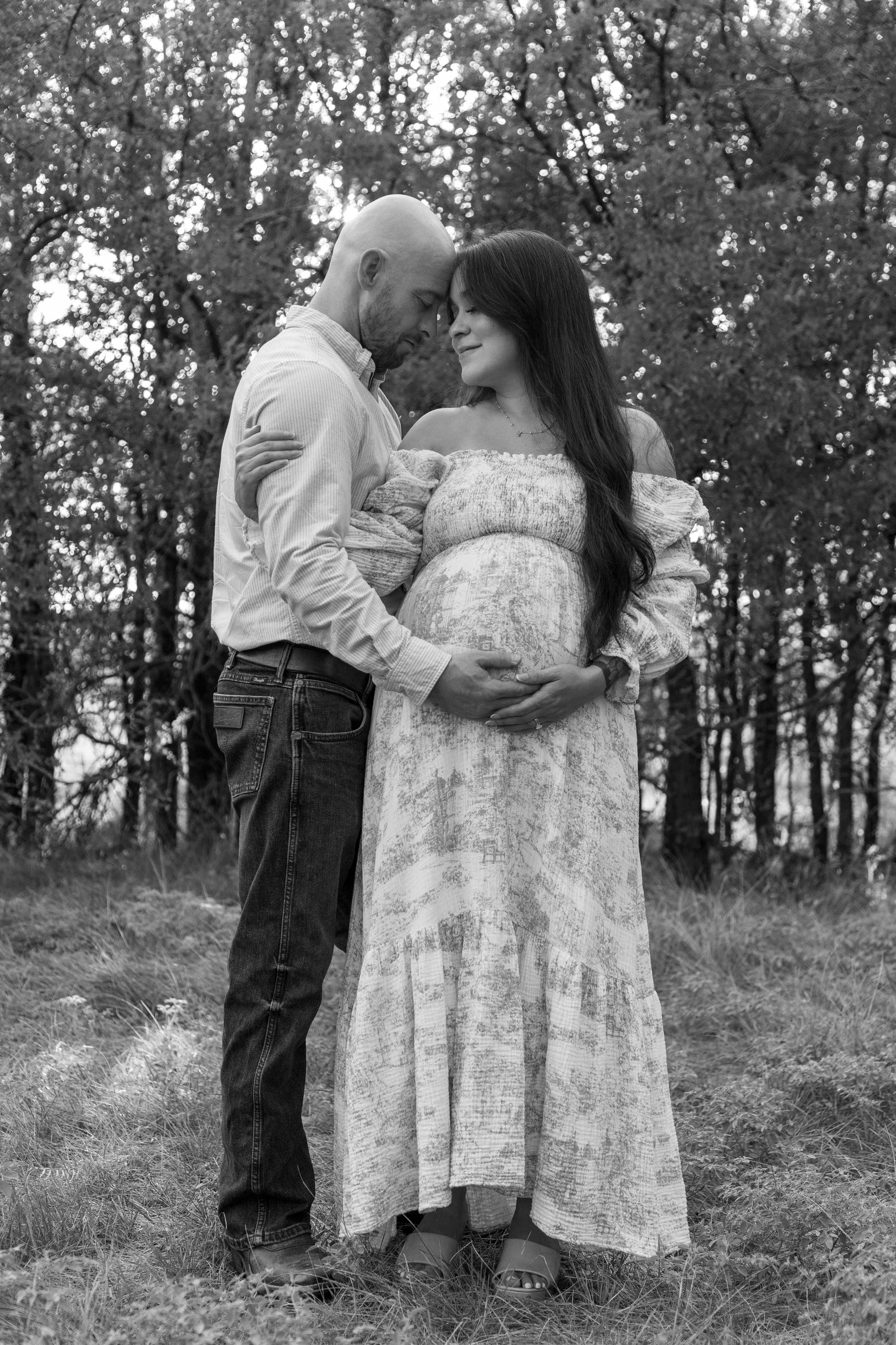 A pregnant woman and a man stand outdoors in a wooded area, with their foreheads touching and hands on her belly, creating a tender, loving moment.