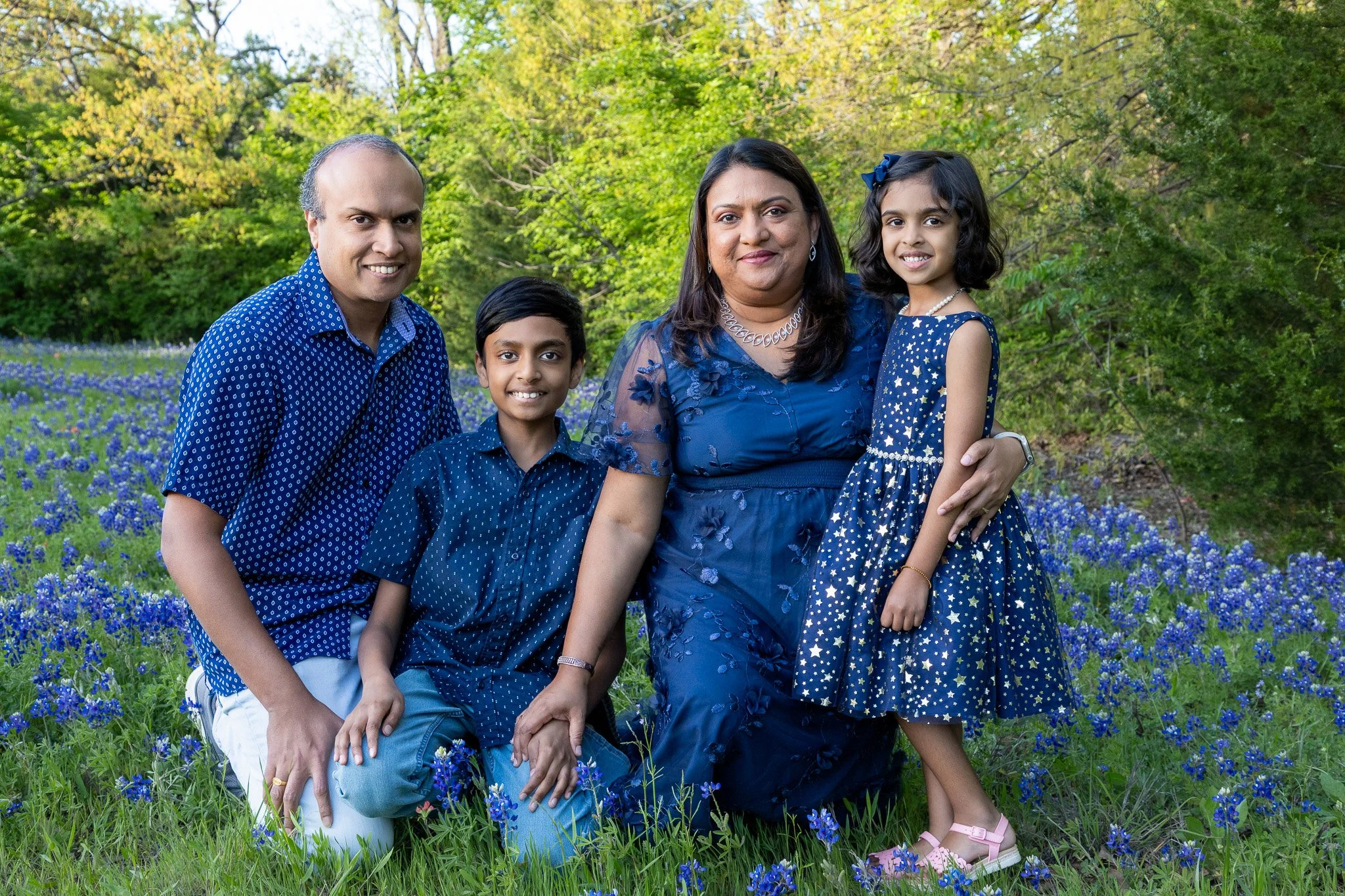 A family of five standing in a field of blue flowers, with green trees in the background, all dressed in blue.