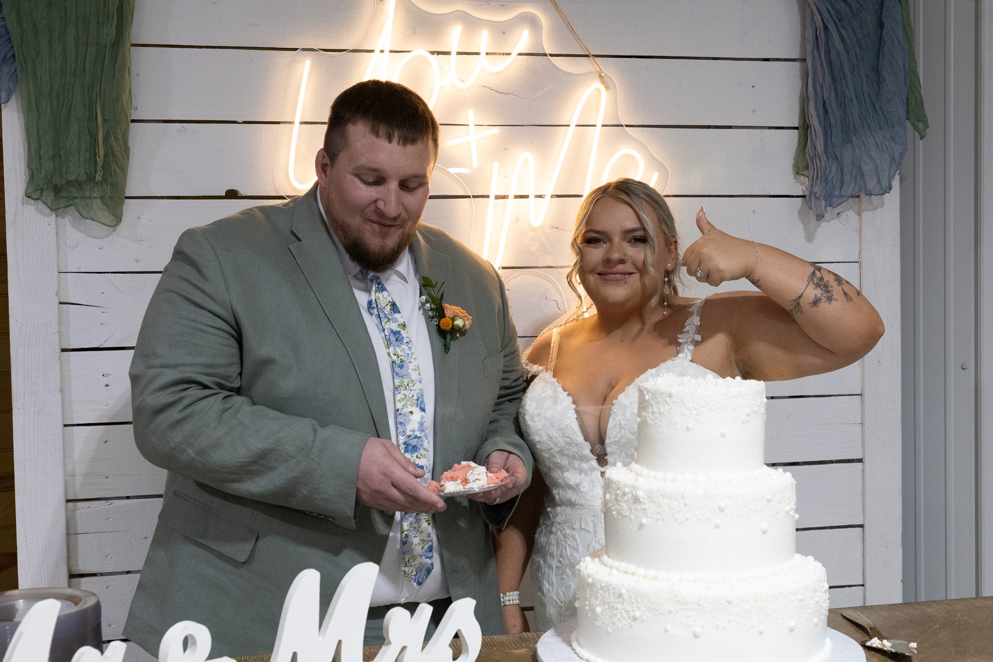A newlywed couple stands behind a three-tier white wedding cake at their wedding reception. The bride is smiling and flexing her arm, showing a tattoo. The groom is holding a plate of cake. They are standing in front of a white wooden wall with a neo