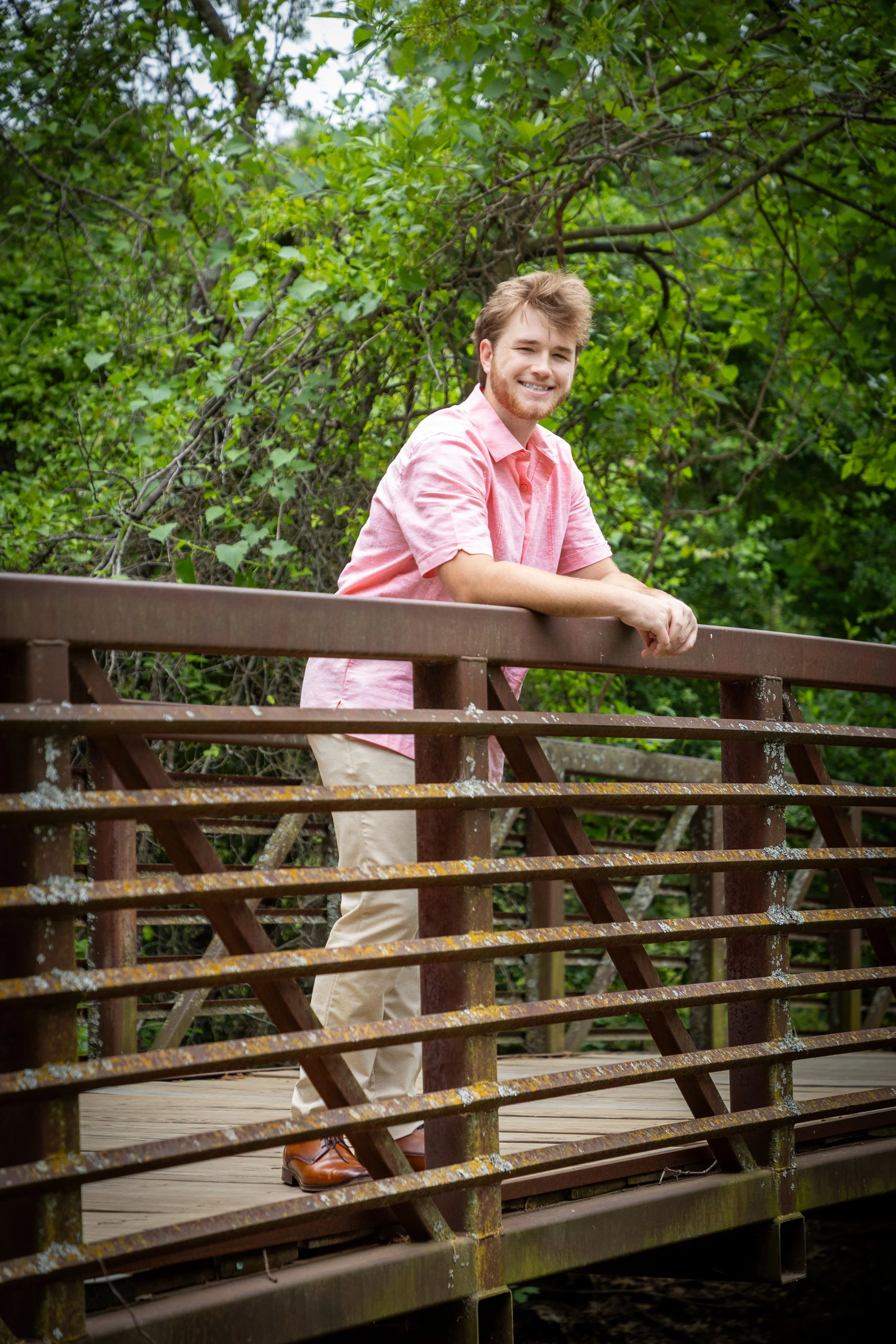 A young man in a pink shirt and beige pants standing on a wooden bridge with metal railings, surrounded by green trees.