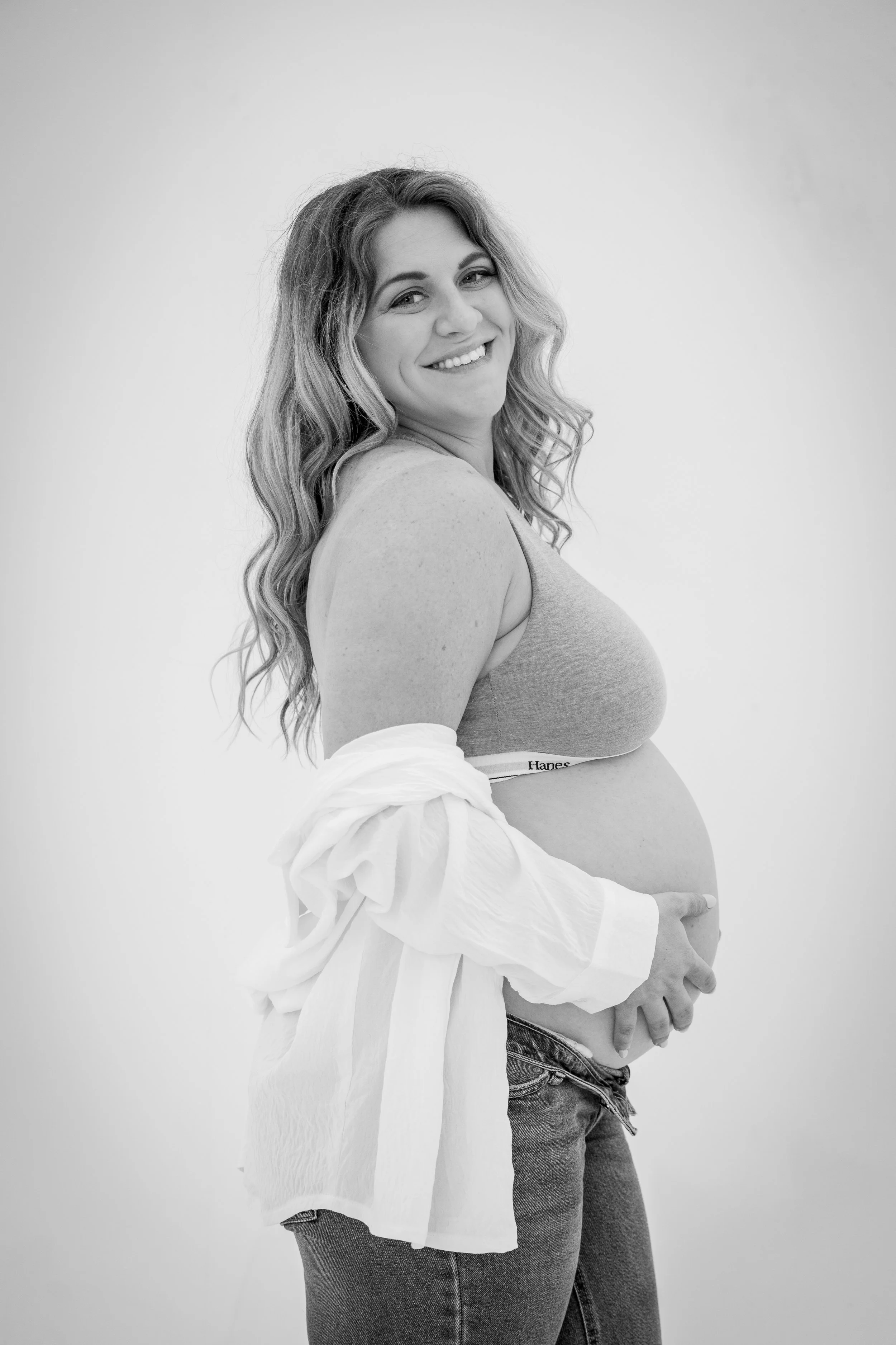 Black and white photo of a pregnant woman with long wavy hair, smiling, holding her belly, wearing a tank top and jeans, with a white shirt draped over her arm.