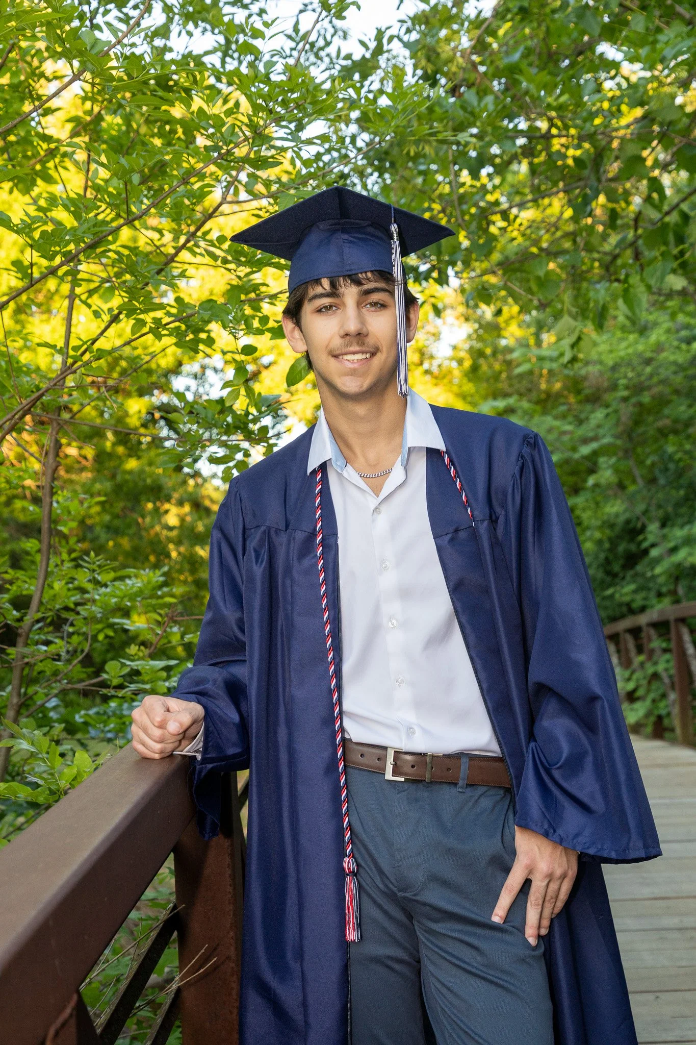 Young man in a blue graduation cap and gown standing outdoors on a wooden bridge surrounded by green trees.