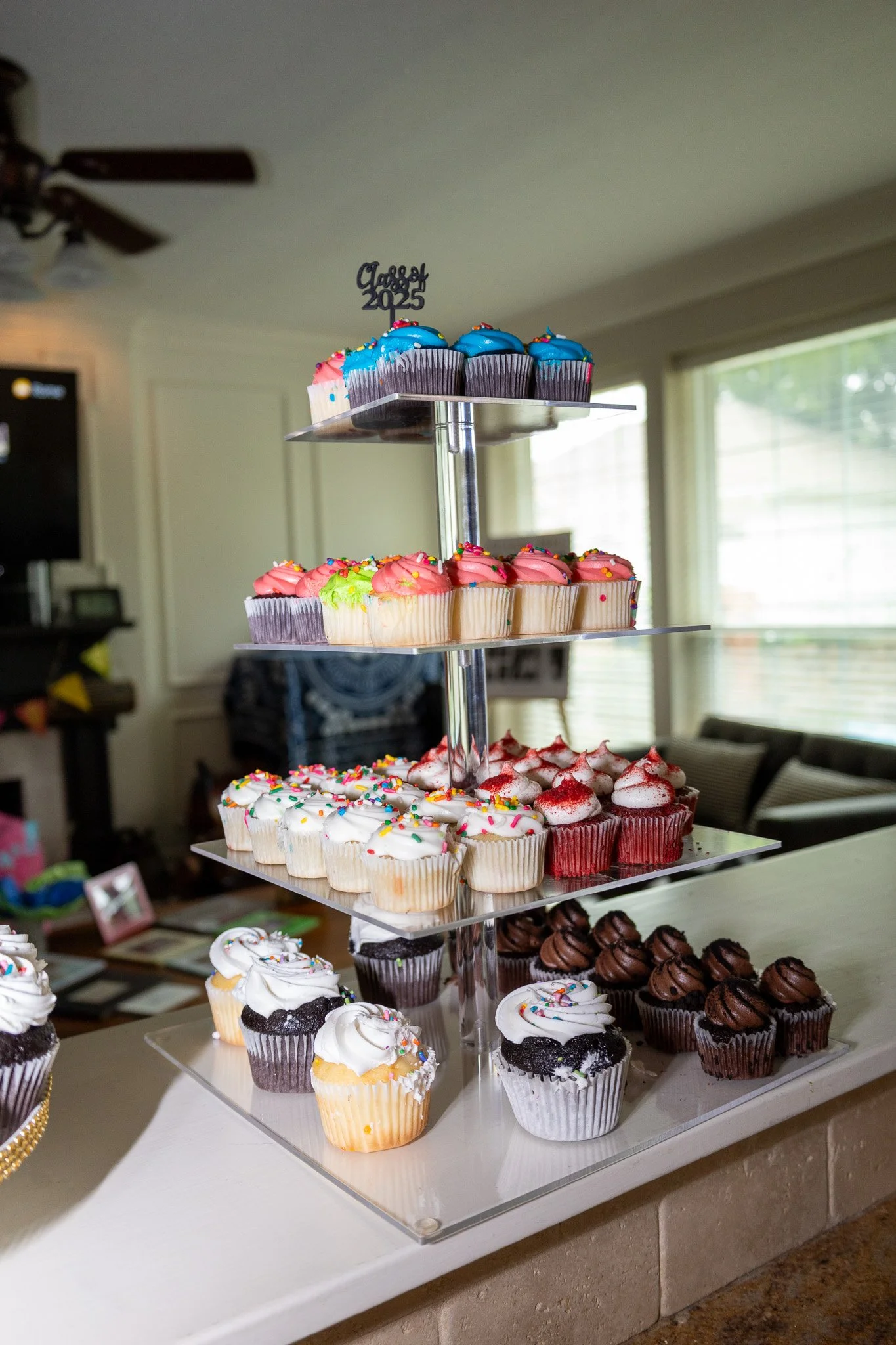 A multi-tiered cupcake display with colorful decorated cupcakes, including red, pink, blue, and chocolate varieties, for a celebration in a home setting.