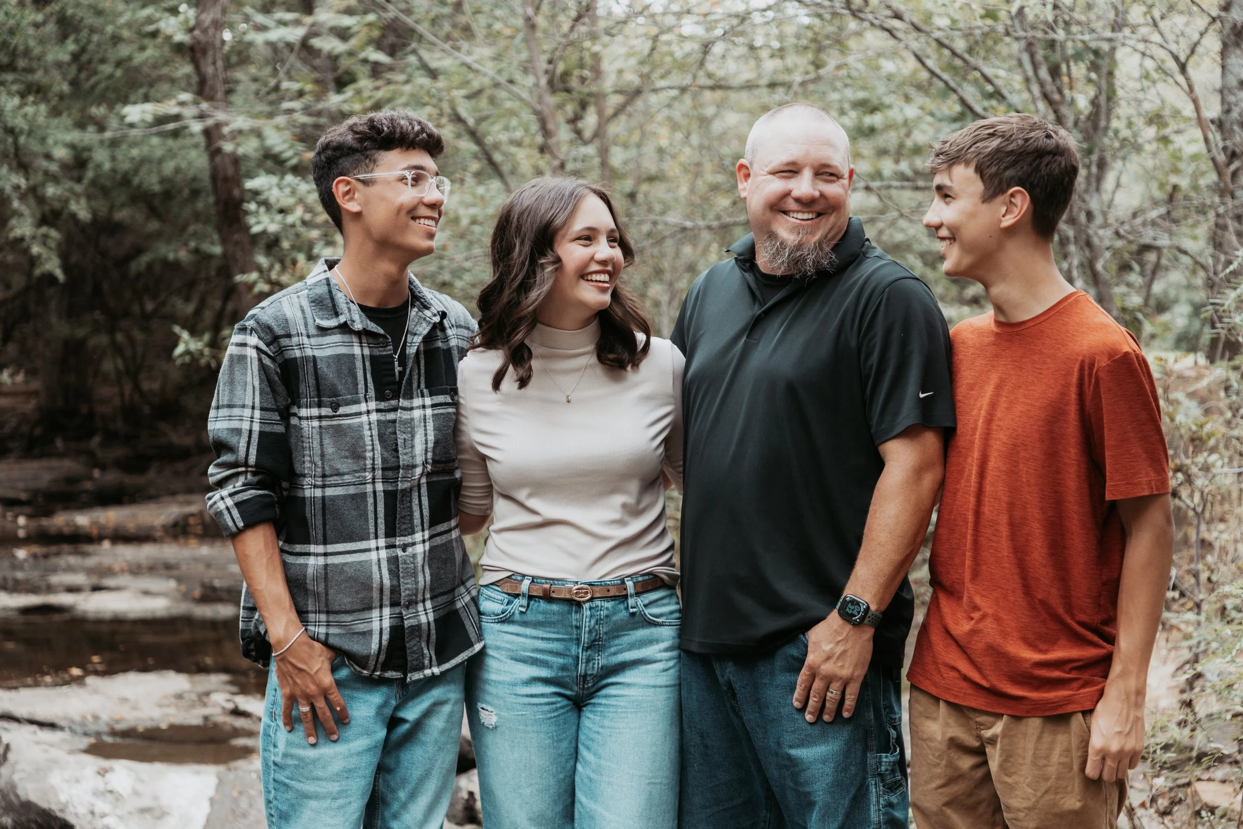 A group of four people standing outdoors in a wooded area, smiling and talking to each other.
