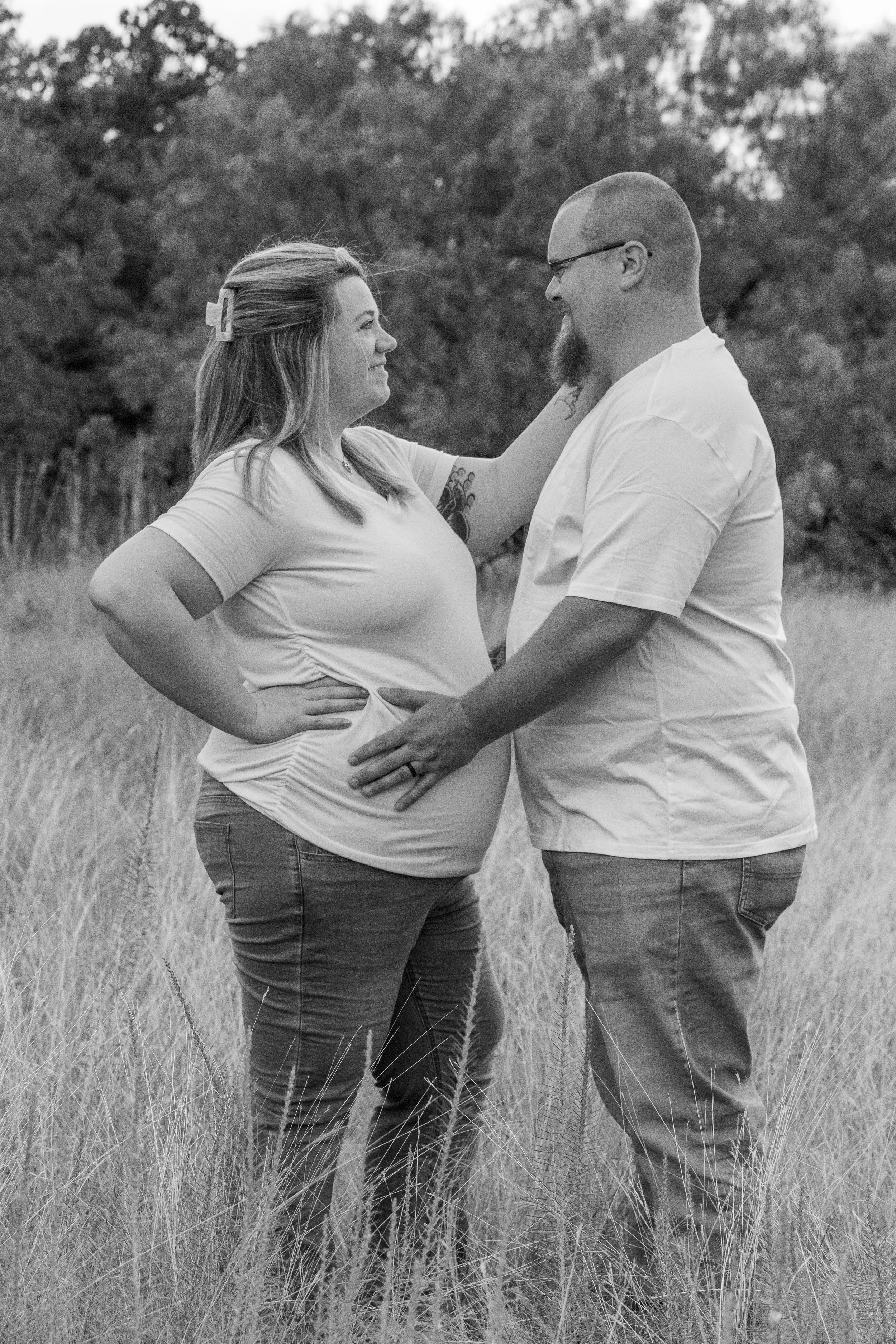 A couple standing in tall grass outdoors, smiling at each other, with trees in the background. The woman has long hair held back with a clip, and the man is bald with a beard and glasses.