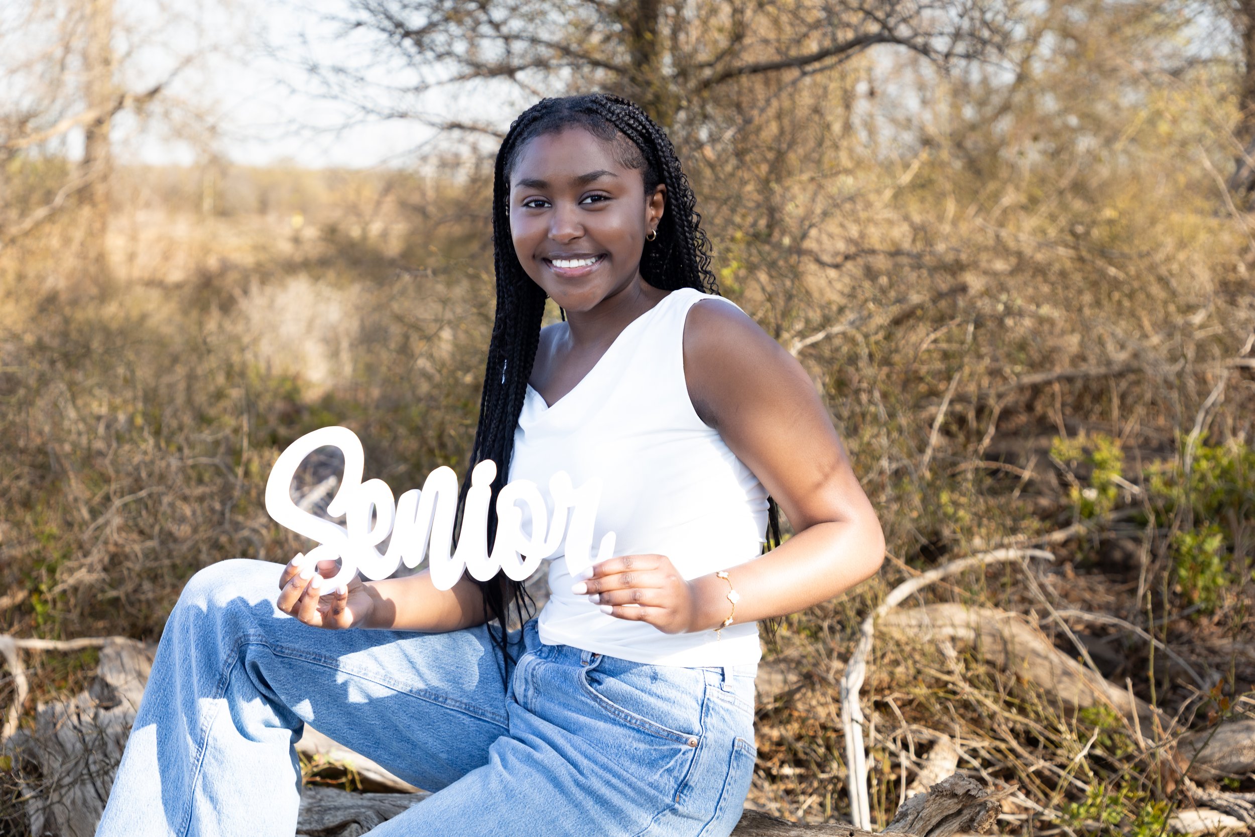 A young woman with braided black hair wearing a white sleeveless top and light blue jeans sitting outdoors on a large rock, smiling and holding a white sign that says 'Smile' in cursive letters. The background features dry trees and bushes in an outd