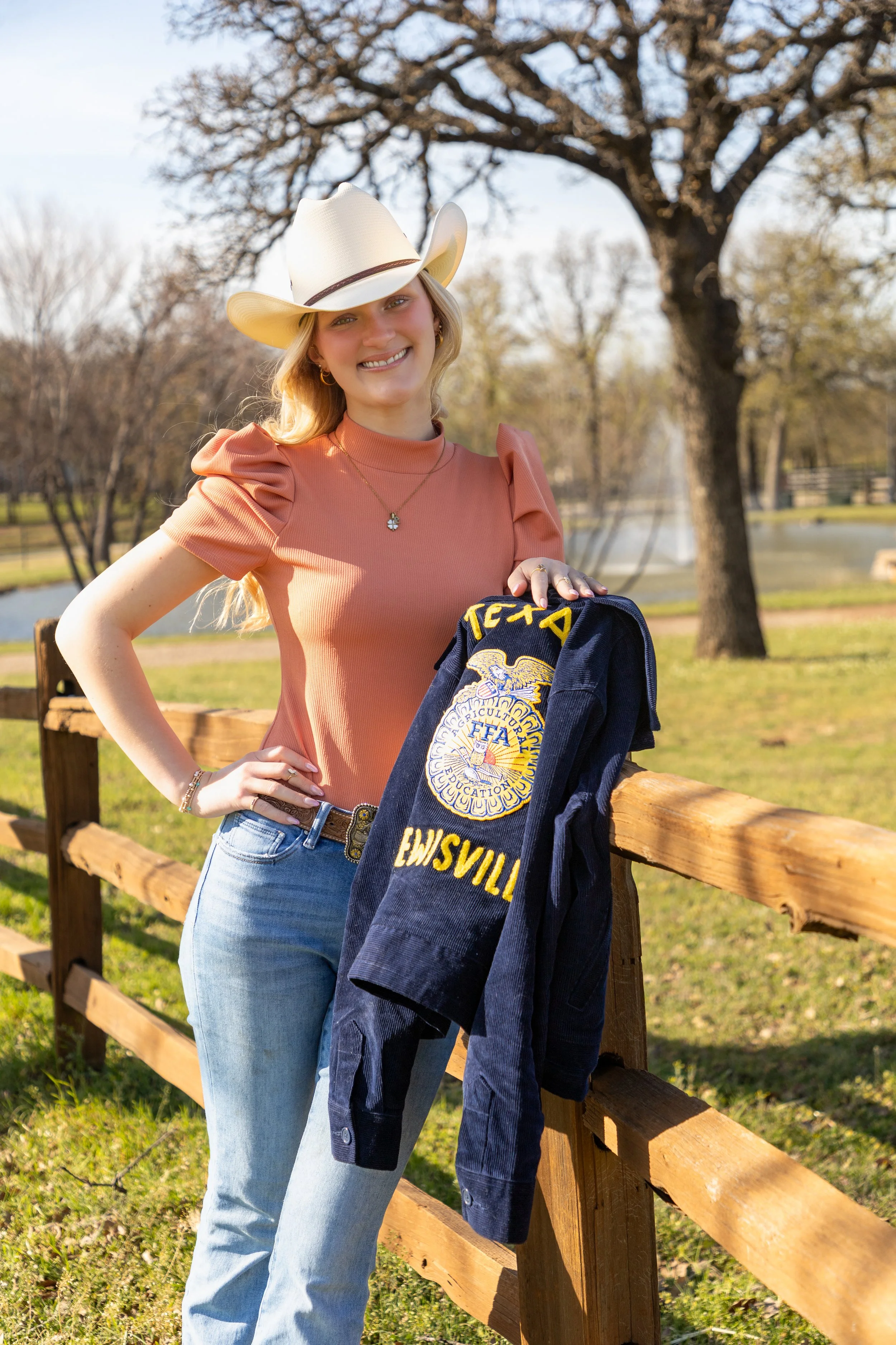 A young woman in a cowboy hat standing outdoors, leaning on a wooden fence, holding a navy blue shirt with an embroidered FFA emblem and the word 'EWSVILLE' on it, with trees and a pond in the background.
